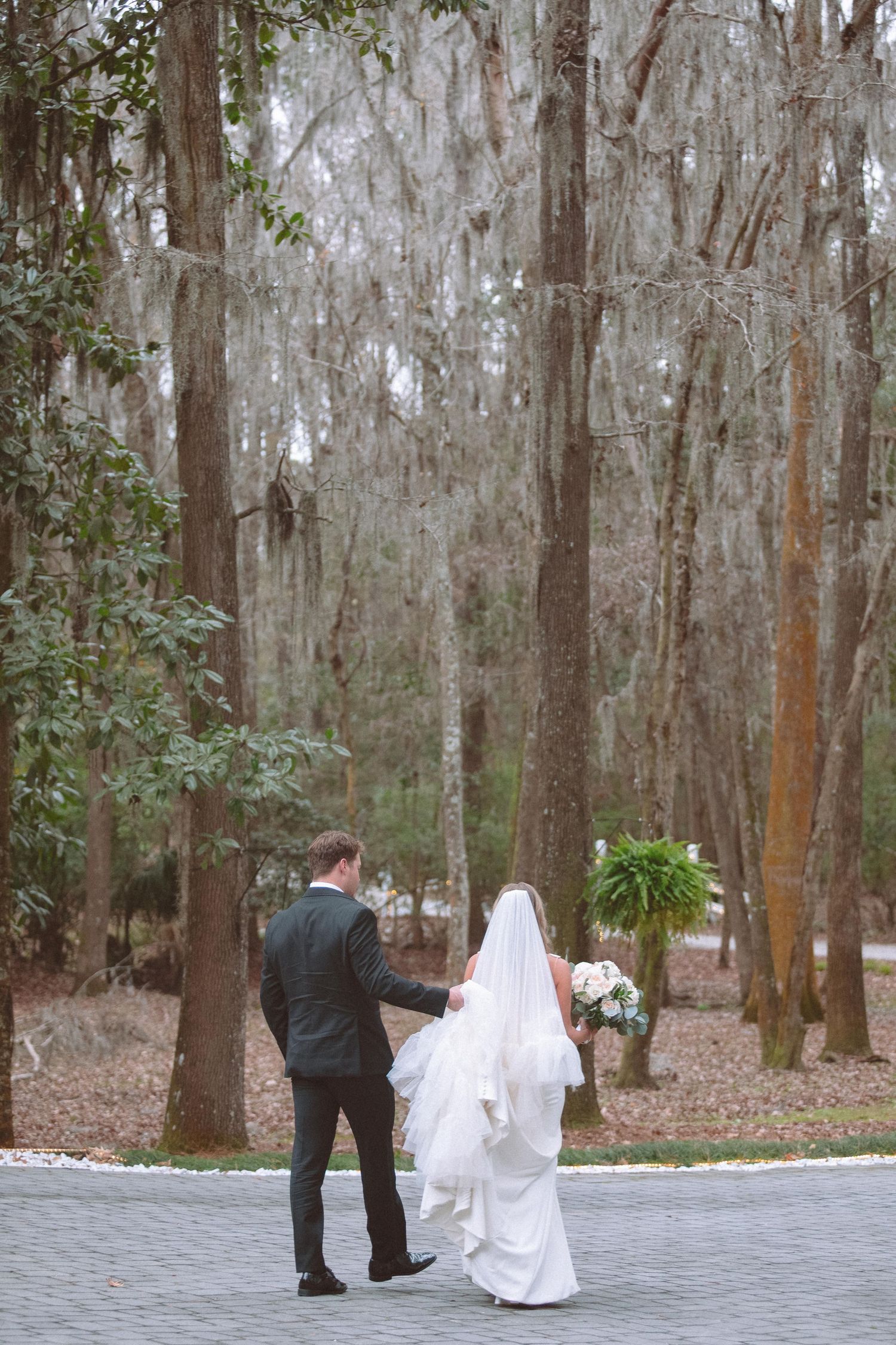 Bride and groom walk hand in hand along tree-lined path draped with Spanish moss.