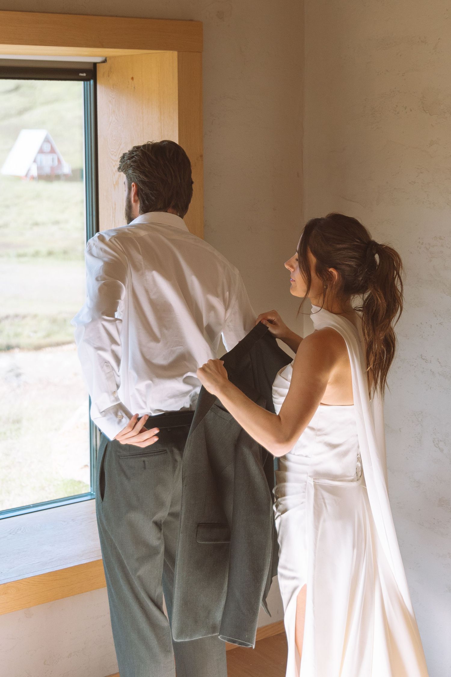 Bride helping groom into his gray suit jacket while standing by a large window.