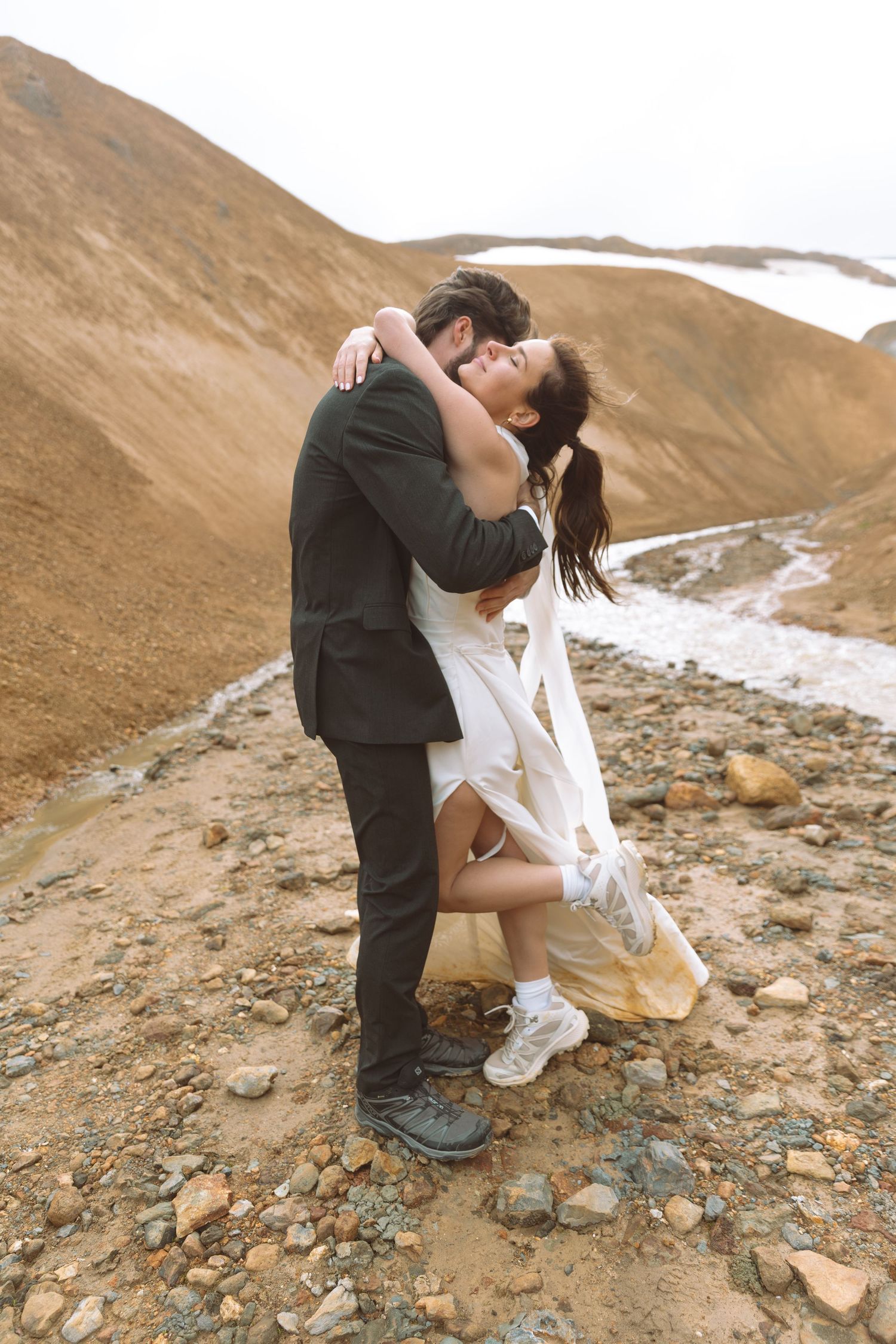 Couple shares a passionate kiss on a rocky hillside, bride in white dress and groom in dark suit.