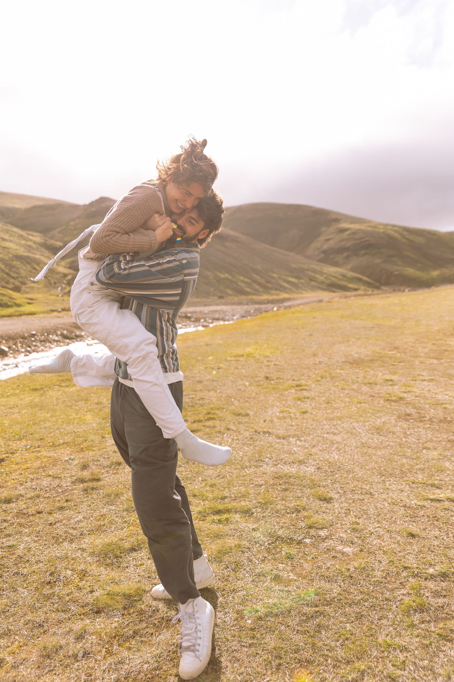 Man giving woman a piggyback ride on a sunny green hillside near a stream in Iceland.