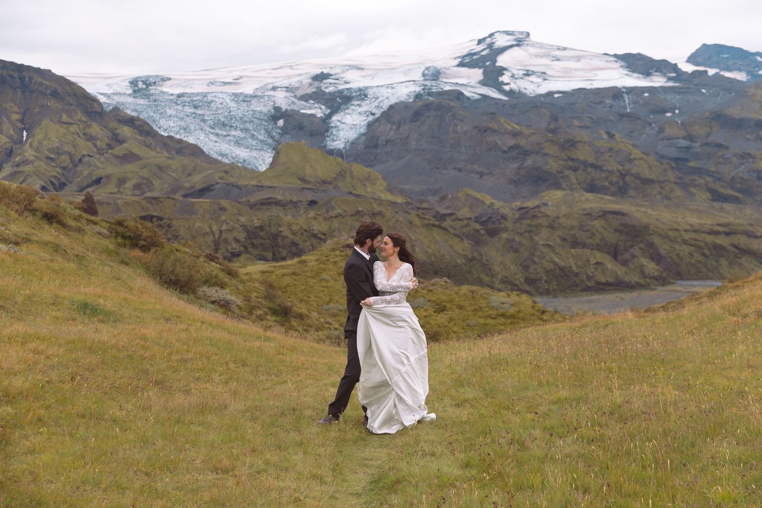 Couple sharing a kiss on green hillside with breathtaking Icelandic glacier mountain backdrop.