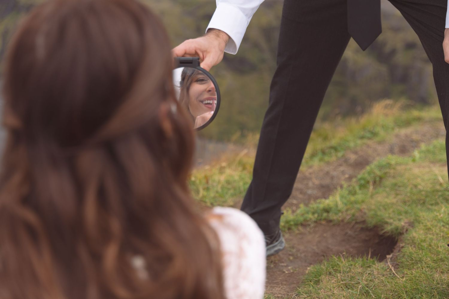 Groom holds round mirror reflecting bride's face during romantic outdoor wedding photoshoot on green hillside.