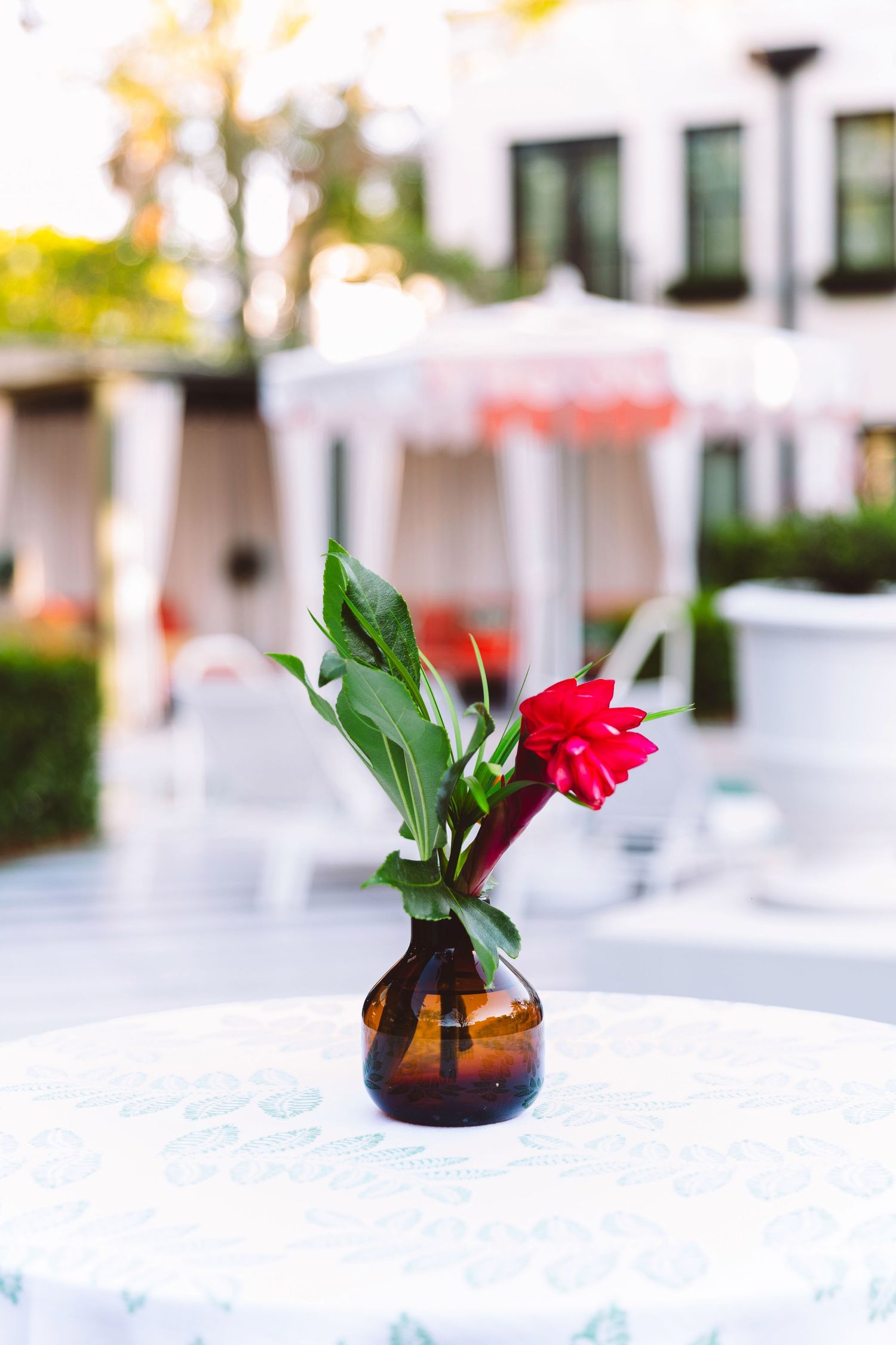 Red tropical flower in amber glass vase on white tablecloth table at outdoor hotel patio setting.