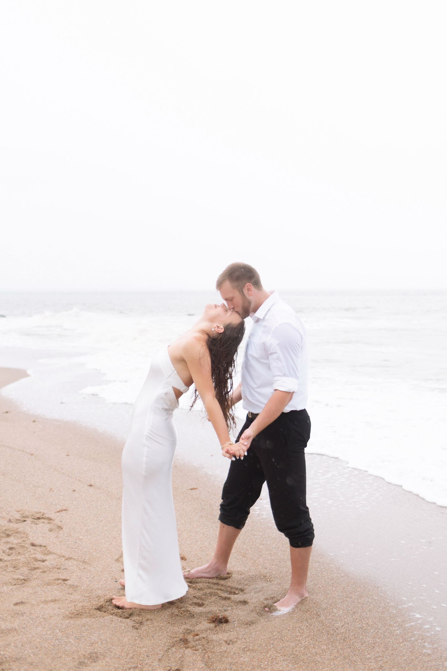 A couple in white shares a romantic kiss on a misty beach shoreline.