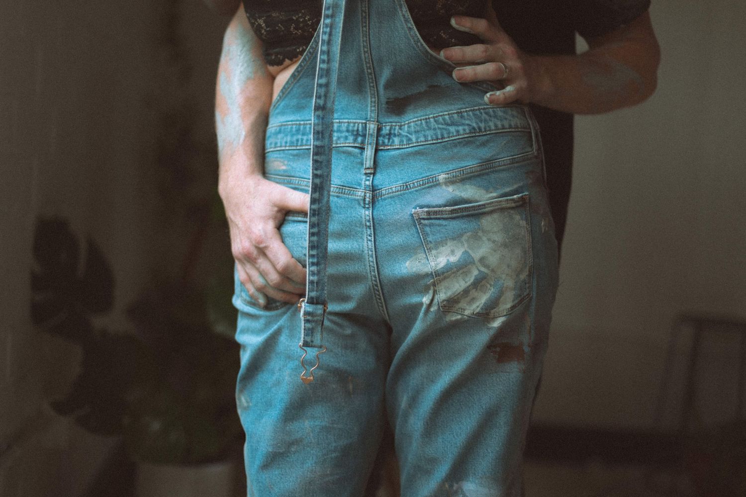 Close-up of person wearing distressed denim overalls with worn patches, standing indoors near plants.