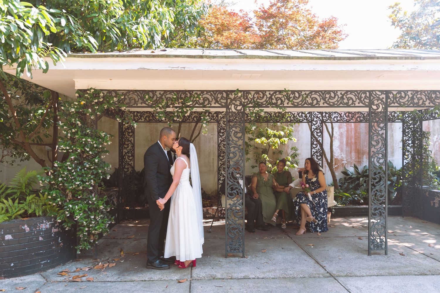 Couple sharing first kiss under decorative iron pergola during intimate outdoor wedding ceremony.