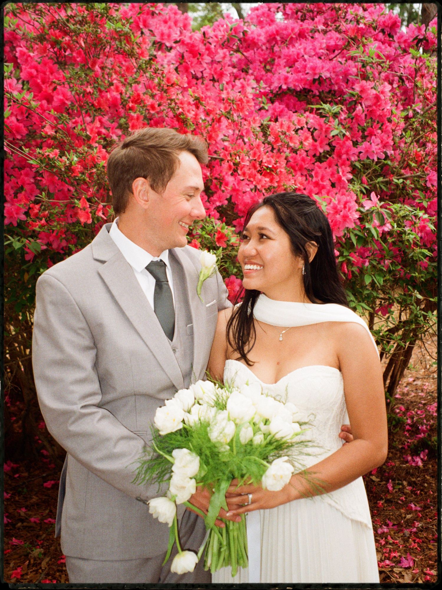 Bride and groom smiling together in front of blooming pink azalea bushes, bride holding white tulips.