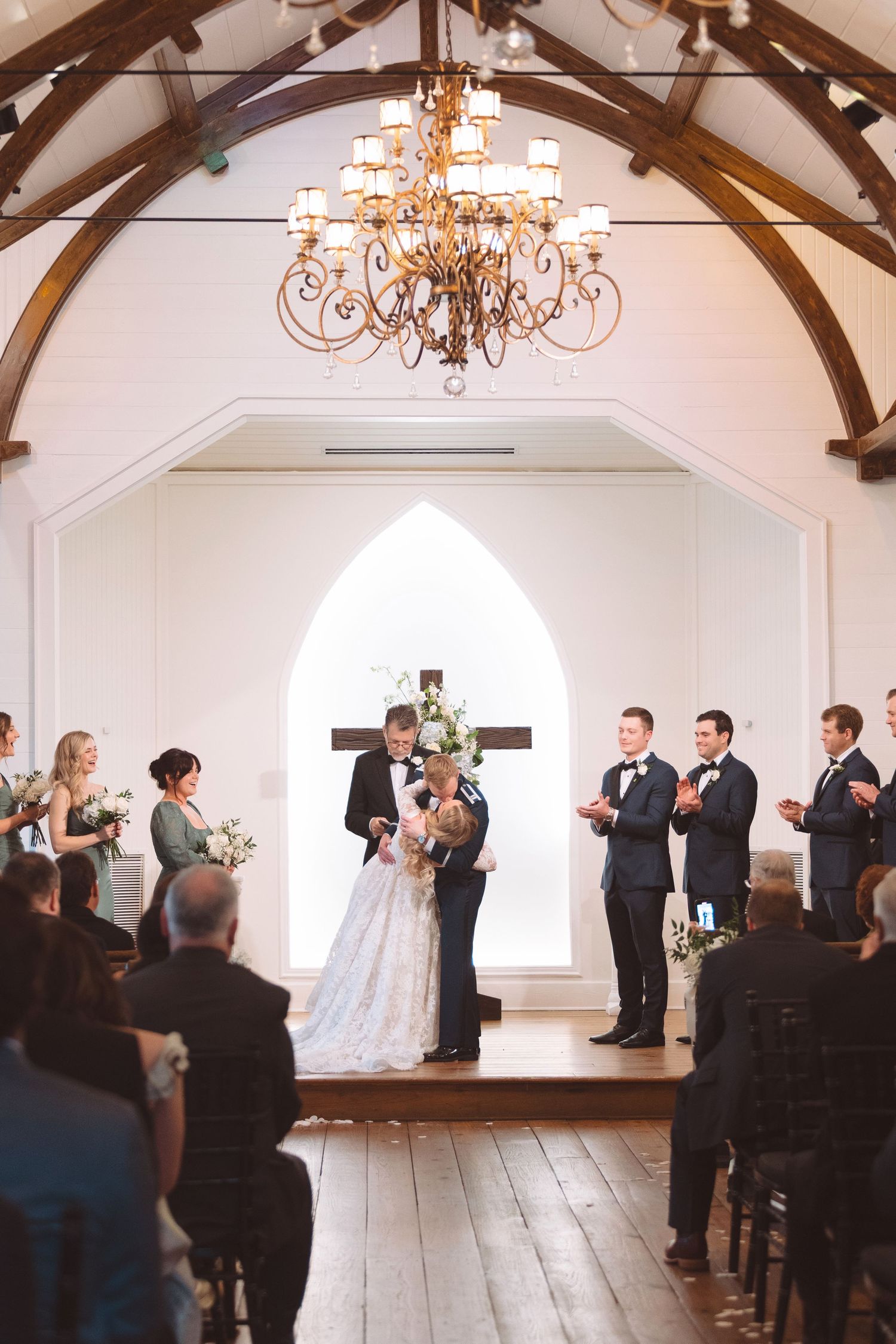 Bride and groom kiss at the altar in a white chapel with arched windows and wood beam ceiling.