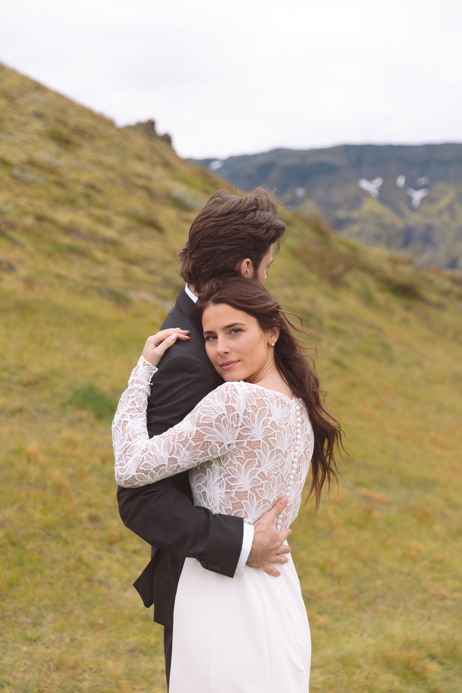Bride in lace gown hugs groom from behind while looking over shoulder at camera outdoors.
