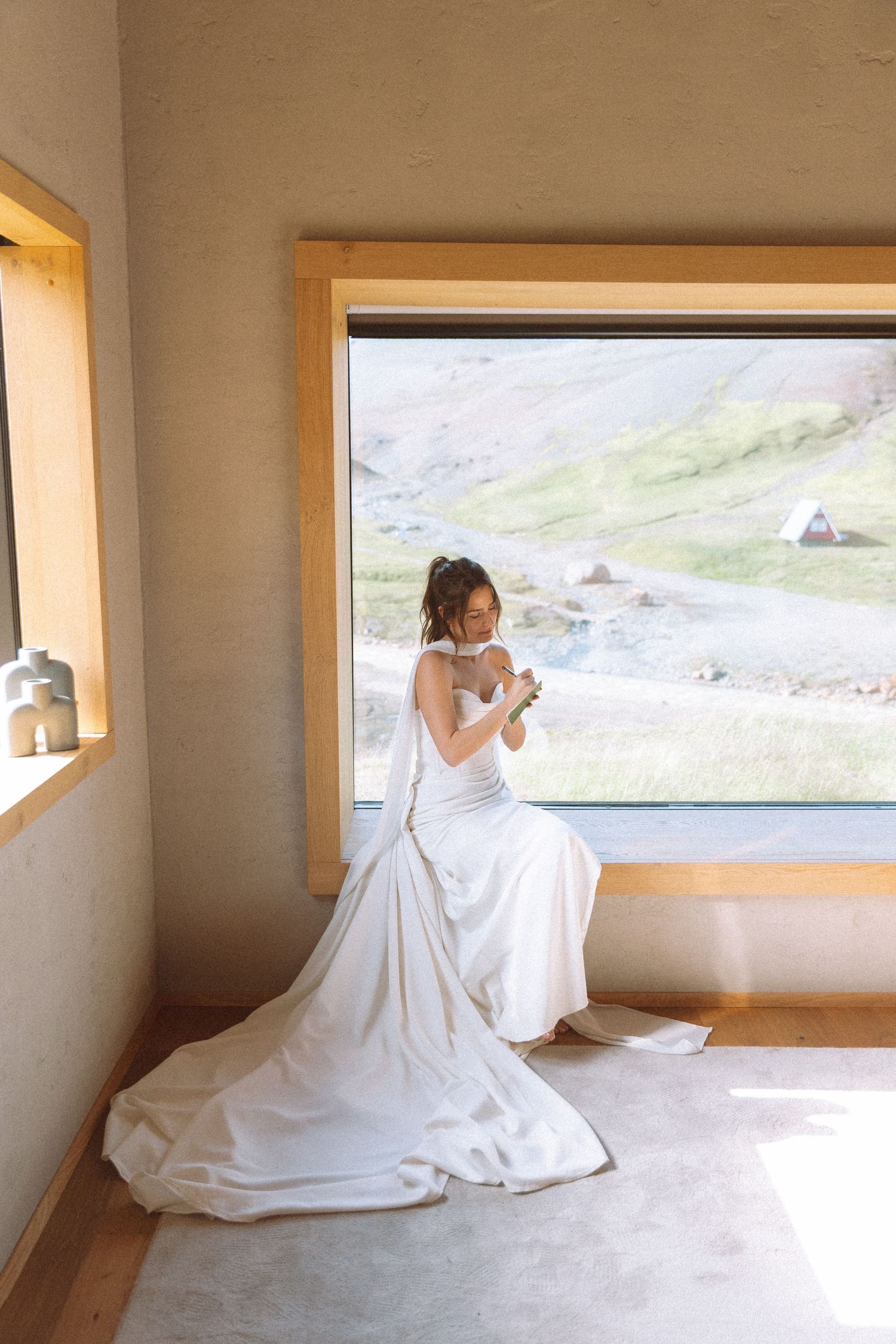 A bride in a white gown with veil sits by a large window with mountain views, holding flowers.
