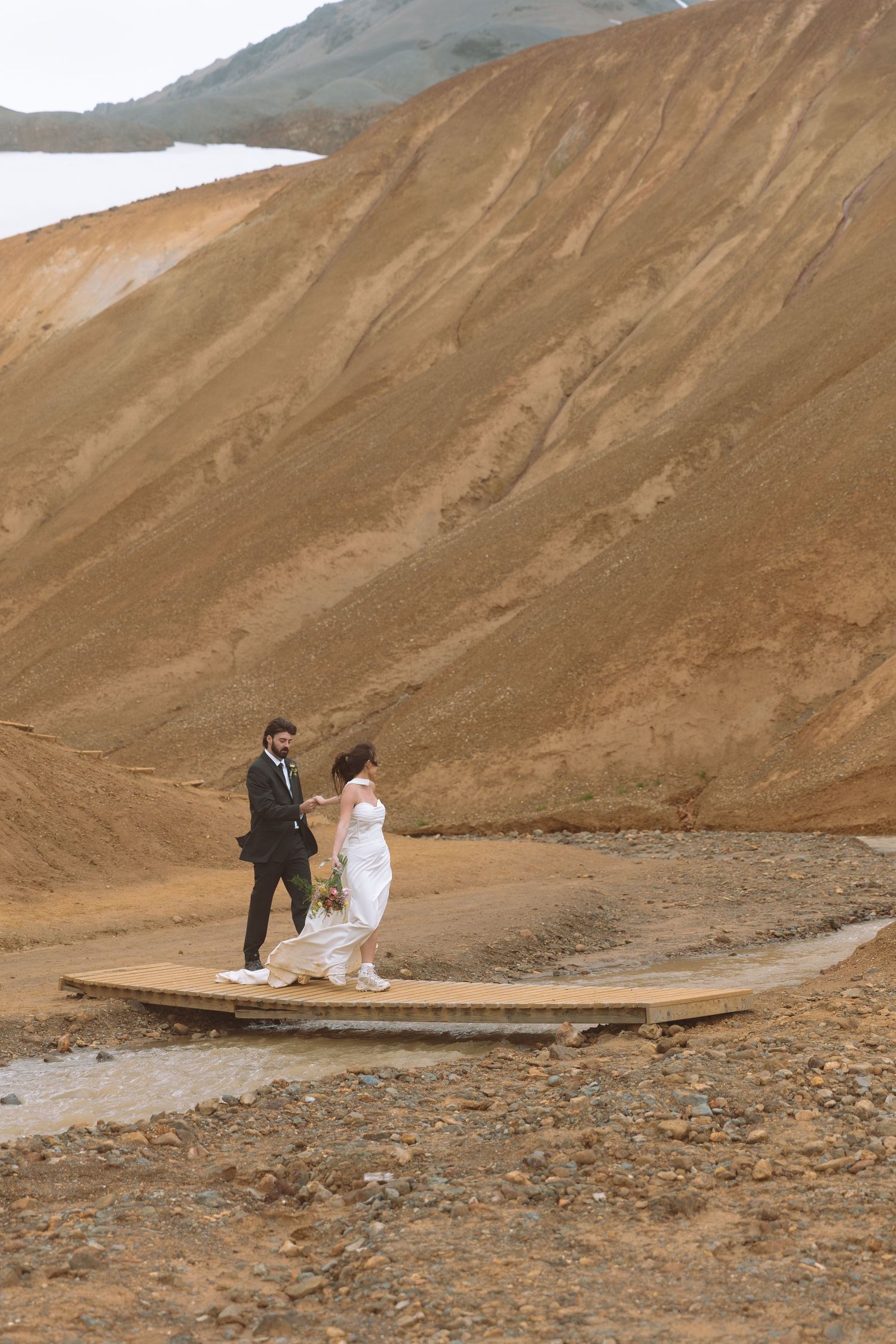 Bride and groom cross a wooden bridge surrounded by vast golden rhyolite mountains in Iceland.