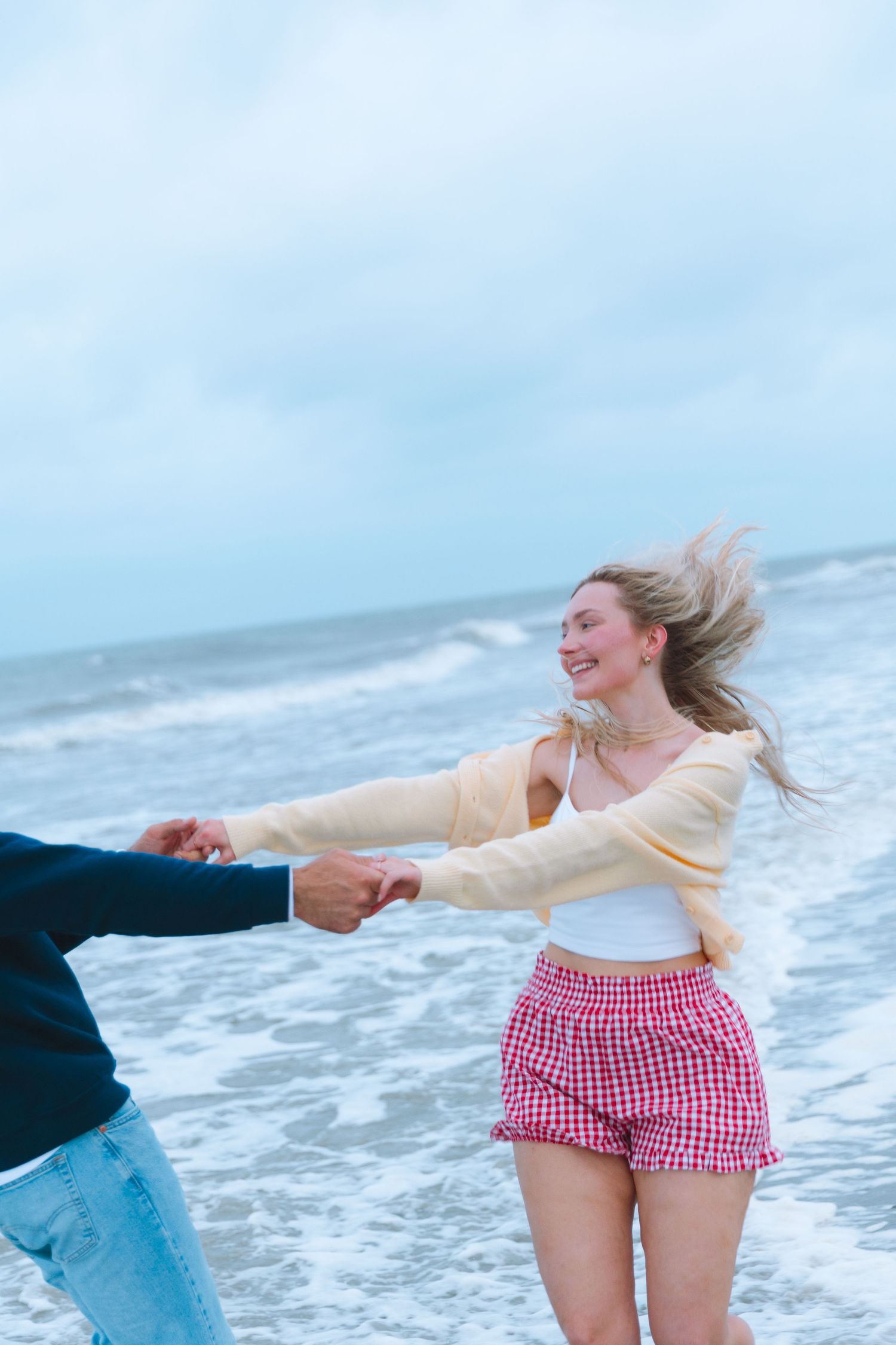 Joyful blonde woman in red plaid shorts and yellow cardigan holding hands with partner on windy beach.