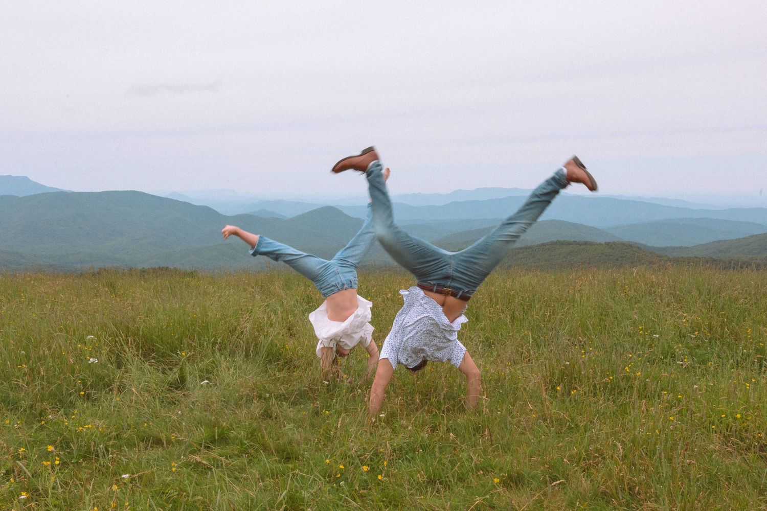 Two people doing simultaneous handstands on a grassy mountain hilltop with scenic valley views.