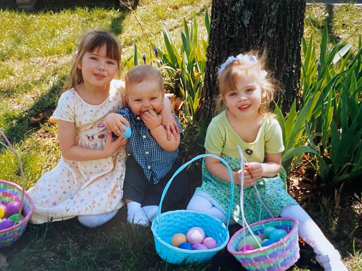 Three young children sitting by a tree with Easter baskets during egg hunt.