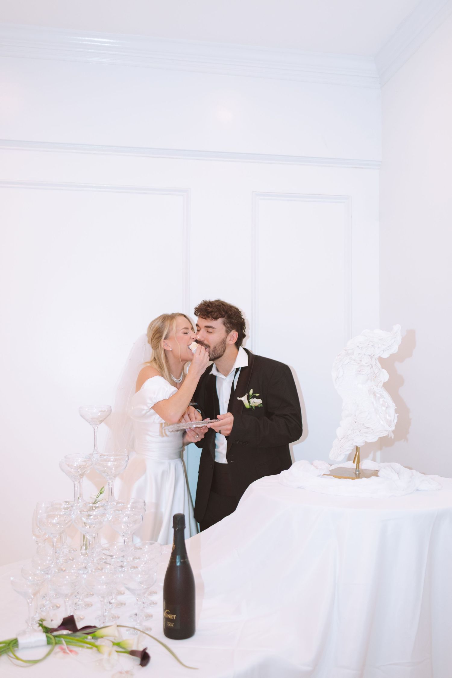 Bride and groom sharing wedding cake at reception table with champagne tower display.