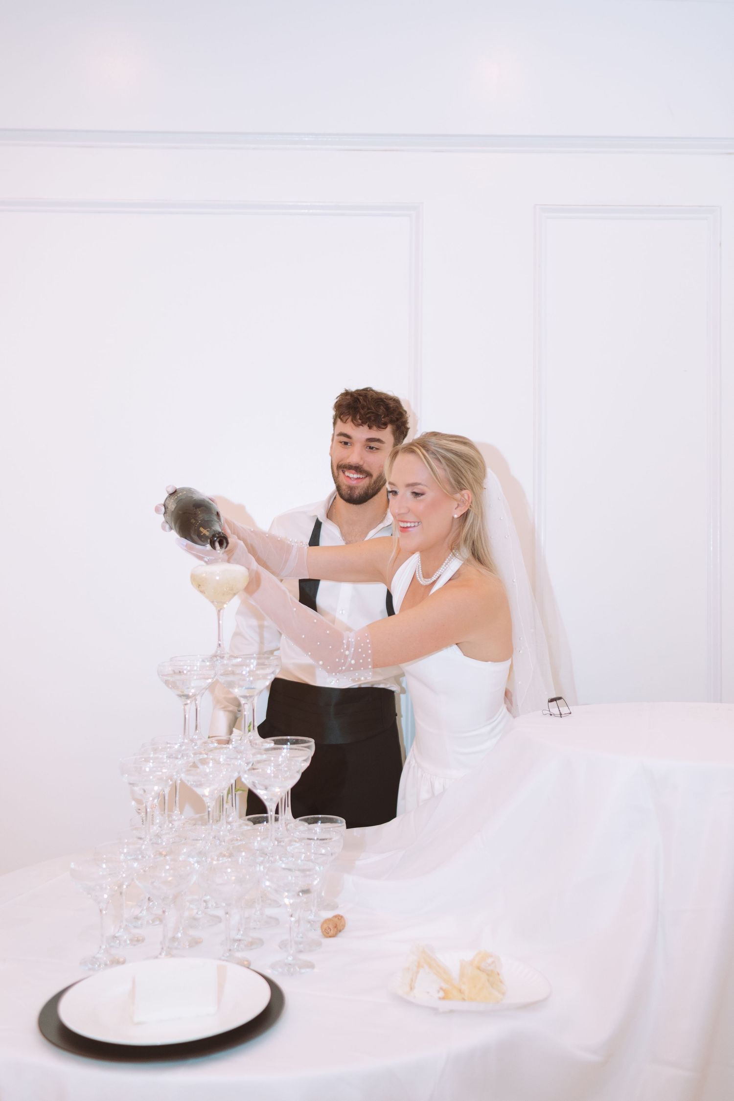 Bride and groom pouring champagne tower at wedding reception in white venue.