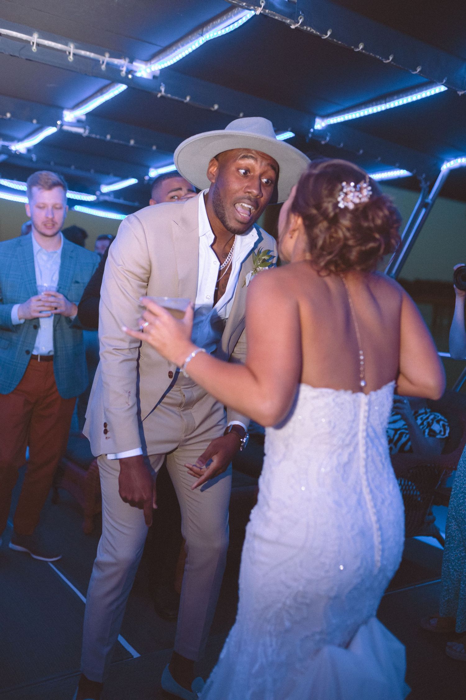 Groom in beige suit and wide-brim hat dances with bride in lace gown at wedding reception.