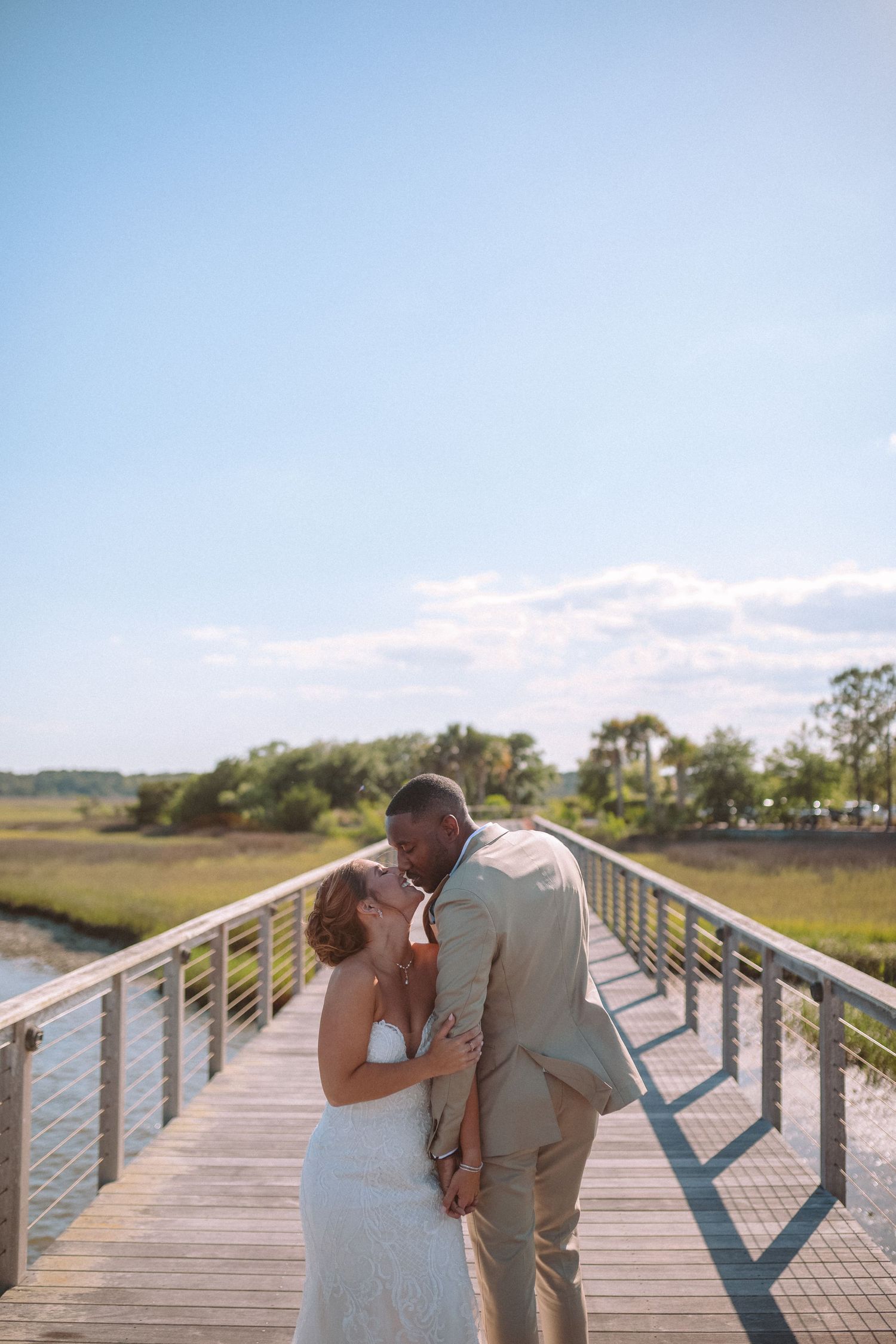 Bride and groom share a kiss on a wooden bridge boardwalk surrounded by marshland under blue sky.