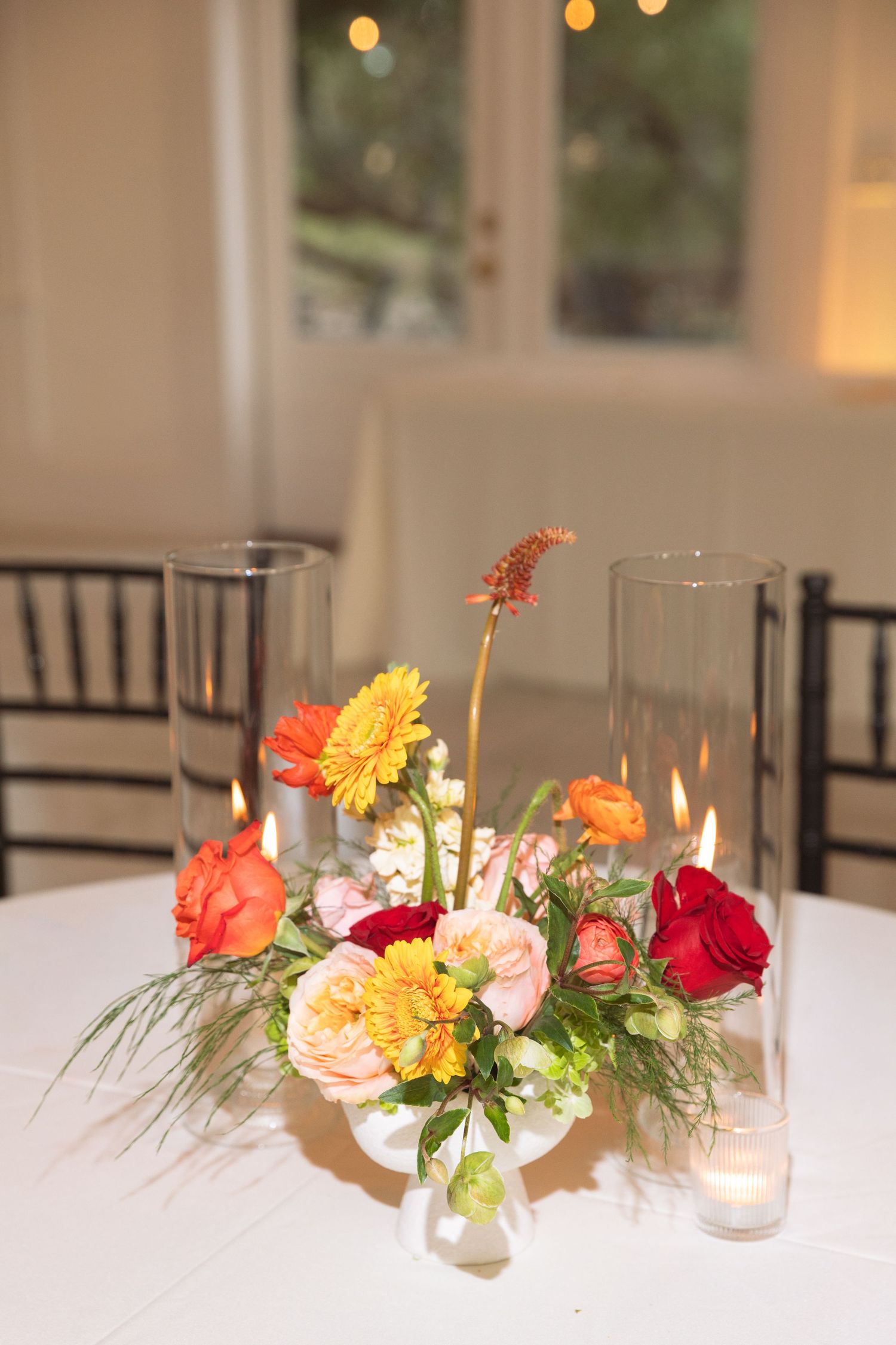 Colorful floral centerpiece with orange gerberas, red roses, and candles in glass cylinders on a white tablecloth.