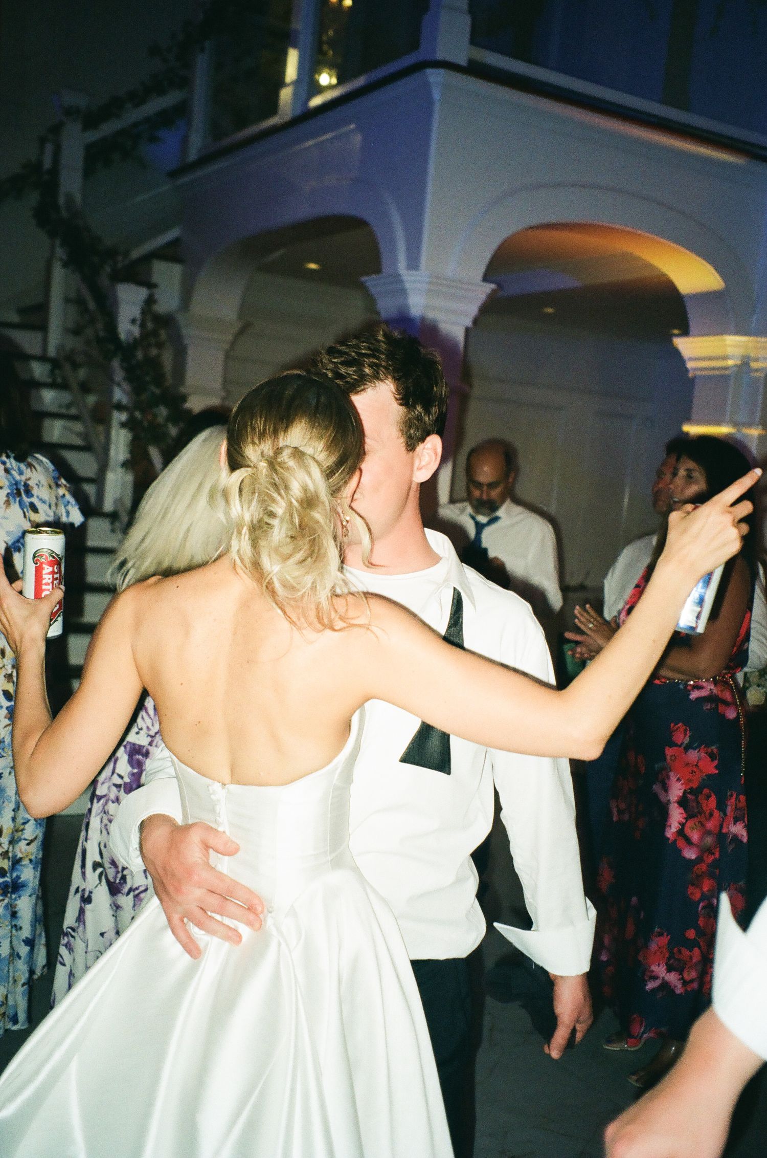 Bride in backless white gown dancing with groom in white jacket at outdoor wedding reception.