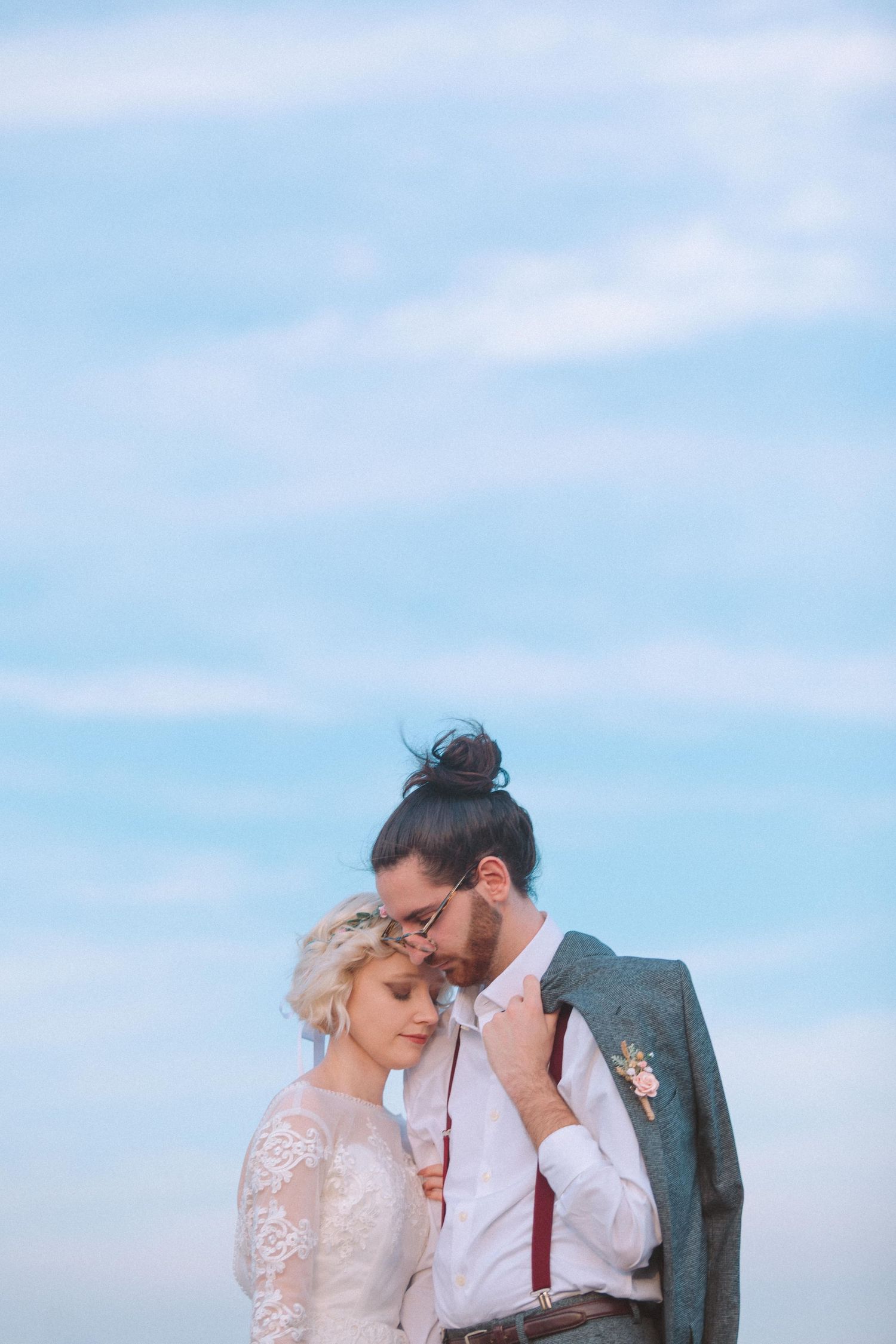 Romantic wedding couple embracing outdoors against a blue sky, groom in suspenders with man bun, bride in lace dress.