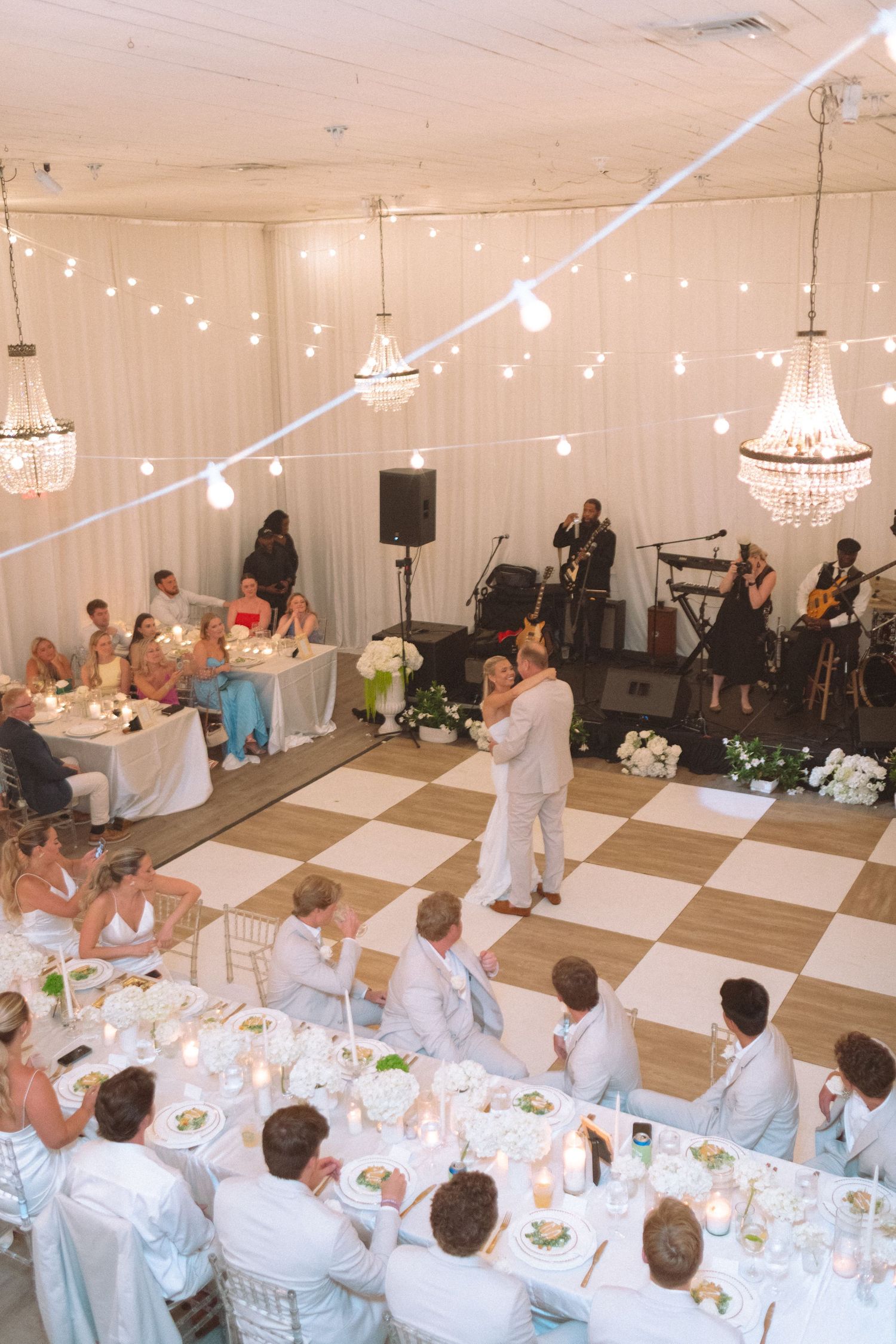 Couple shares first dance on checkered floor under chandeliers in a white draped wedding tent.