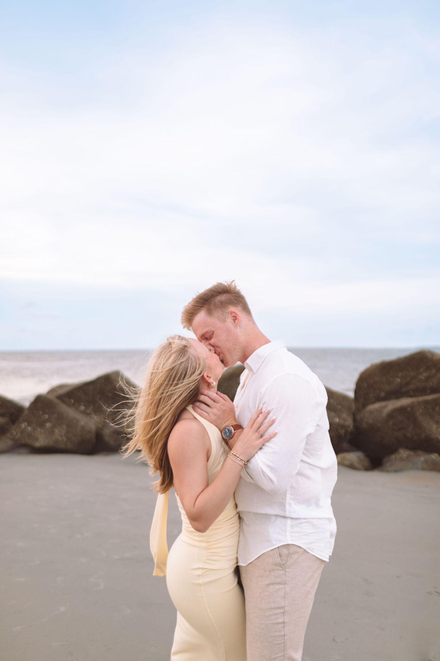A romantic couple kisses on a sandy beach, she in a yellow dress and he in a white shirt with rocks behind.