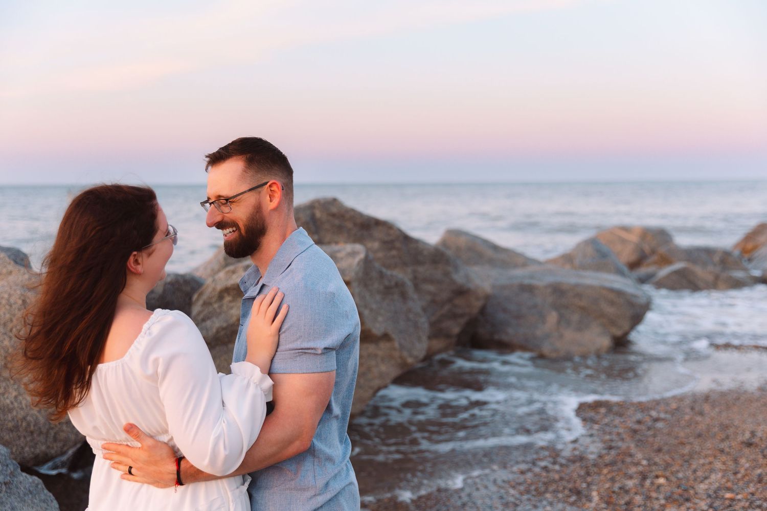 Couple embracing and smiling at each other on rocky beach at dusk near ocean.