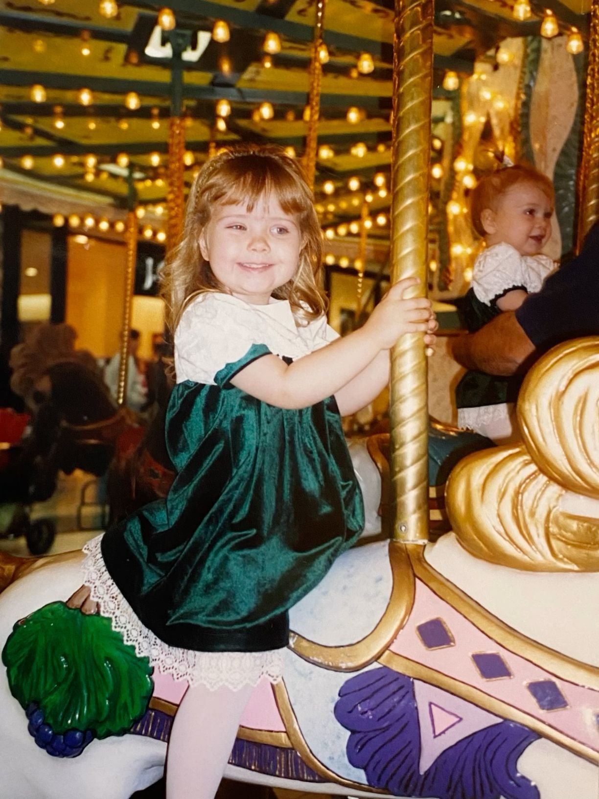 Young girl in green velvet dress smiling on a colorful carousel horse with gold pole and festive lights.