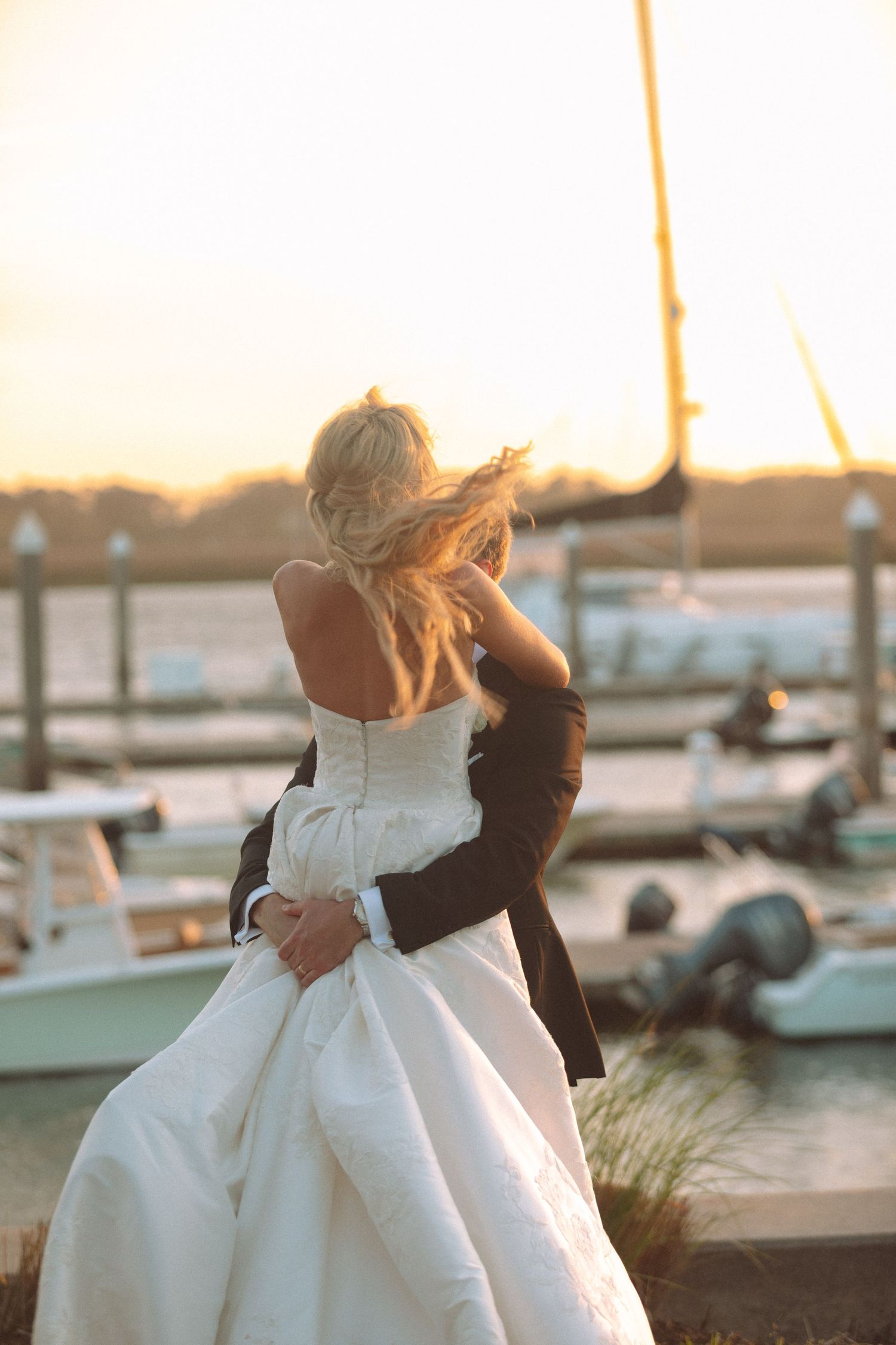 Bride and groom embracing at sunset near a marina, with sailboats and golden light in the background.