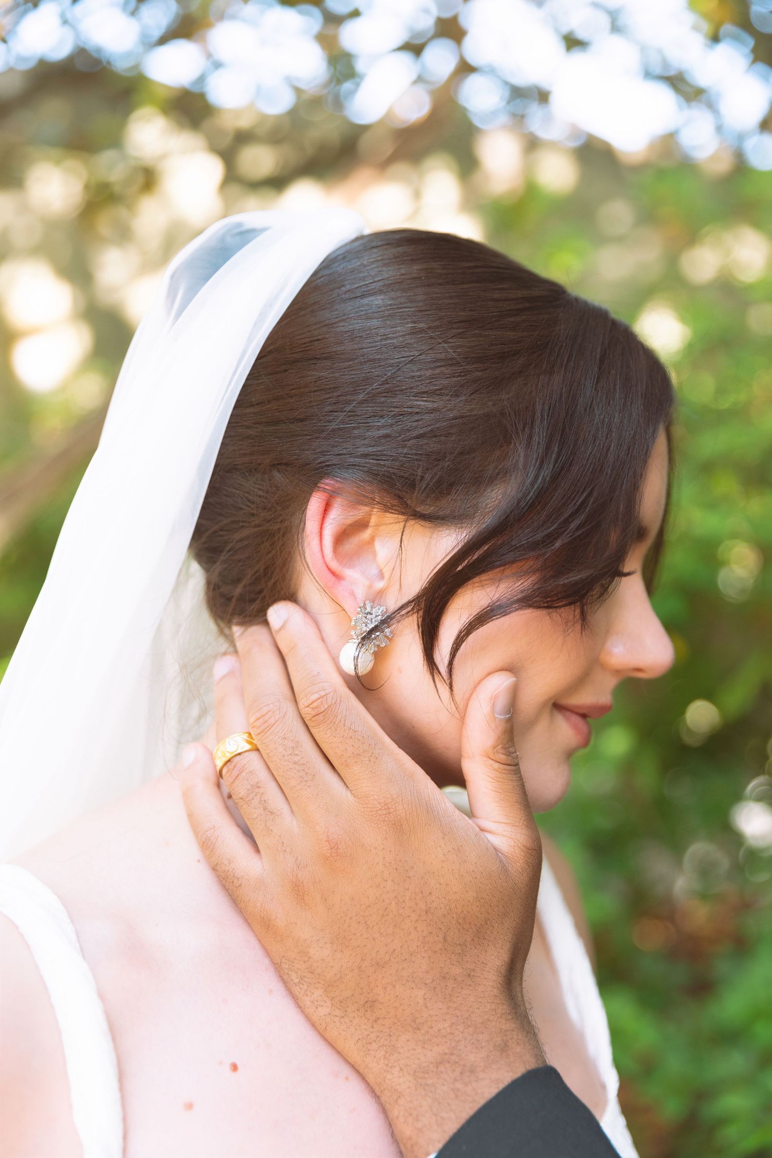 Groom tenderly touches smiling bride's face, showcasing gold wedding ring, as she wears veil and elegant crystal earring.