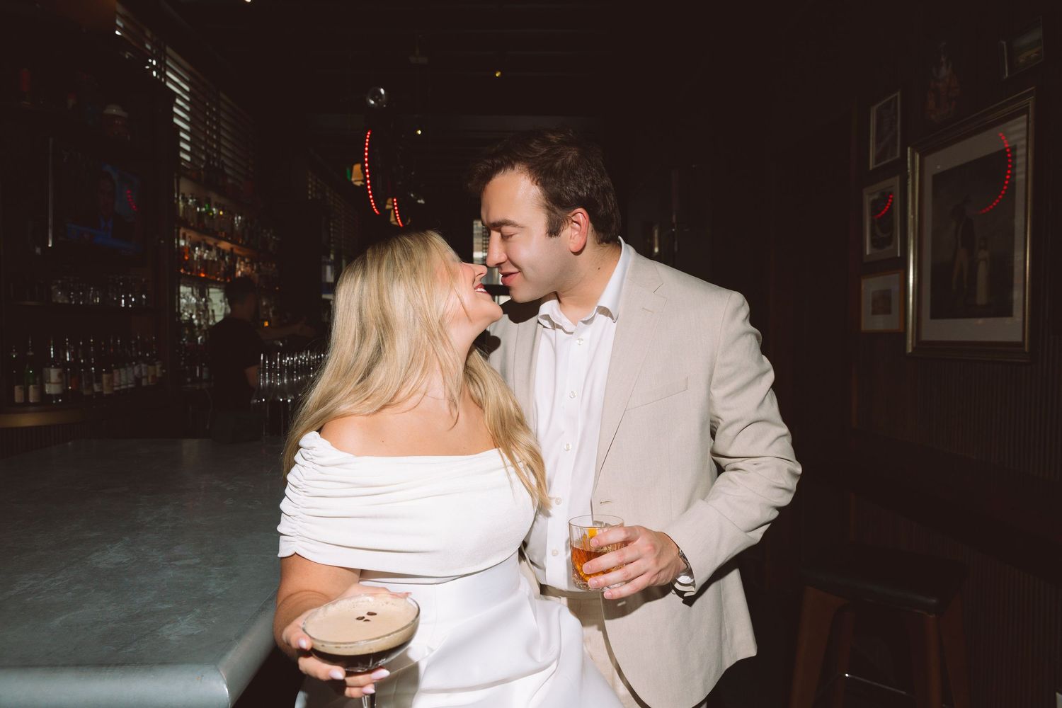 Couple in white outfits sharing a romantic moment at a dimly lit bar, holding cocktails.