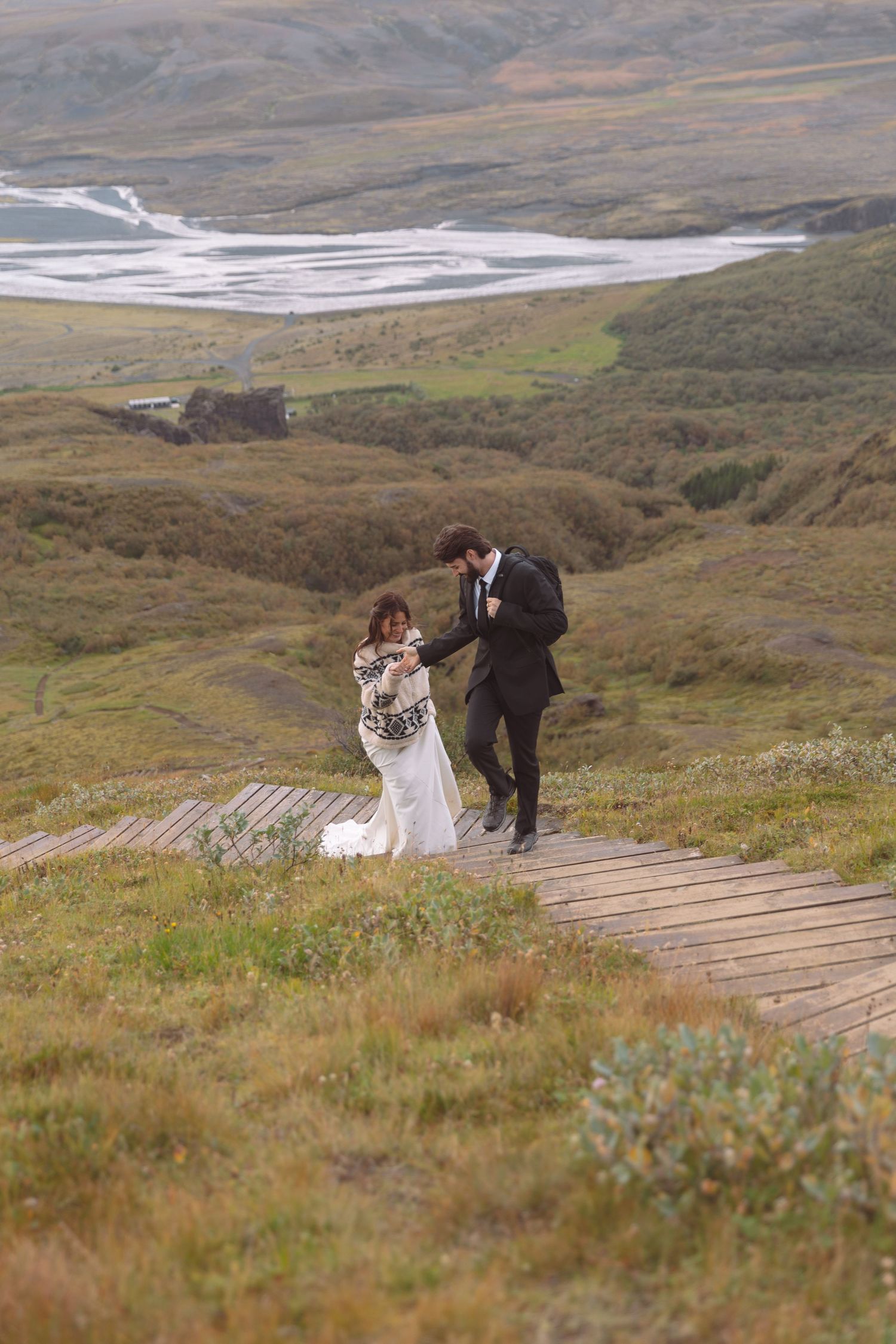 Bride wrapped in cozy sweater and groom walk a wooden boardwalk through Iceland's vast green highlands.