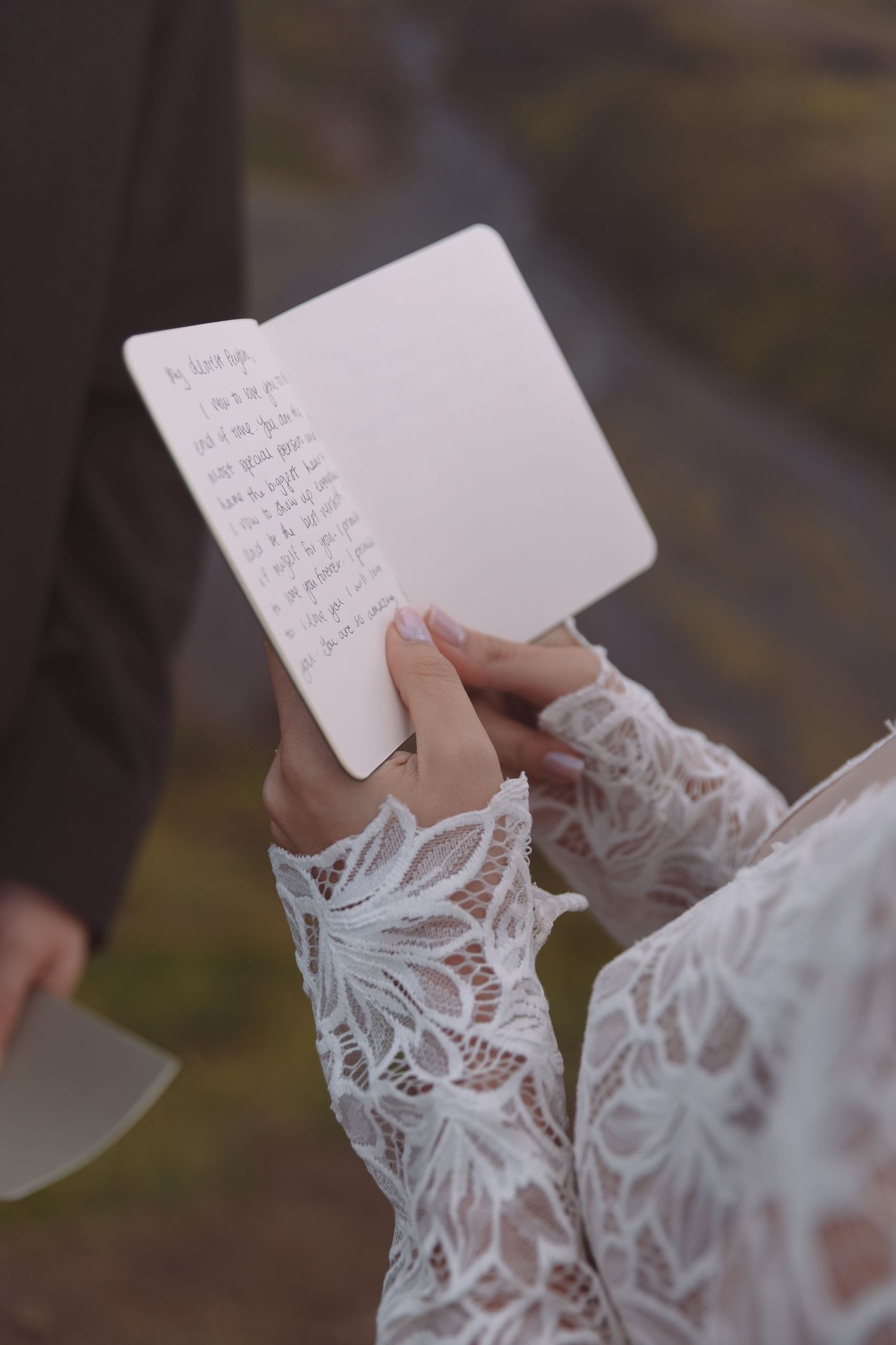 Close-up of bride holding handwritten wedding vows notebook in lace long-sleeve wedding dress outdoors.