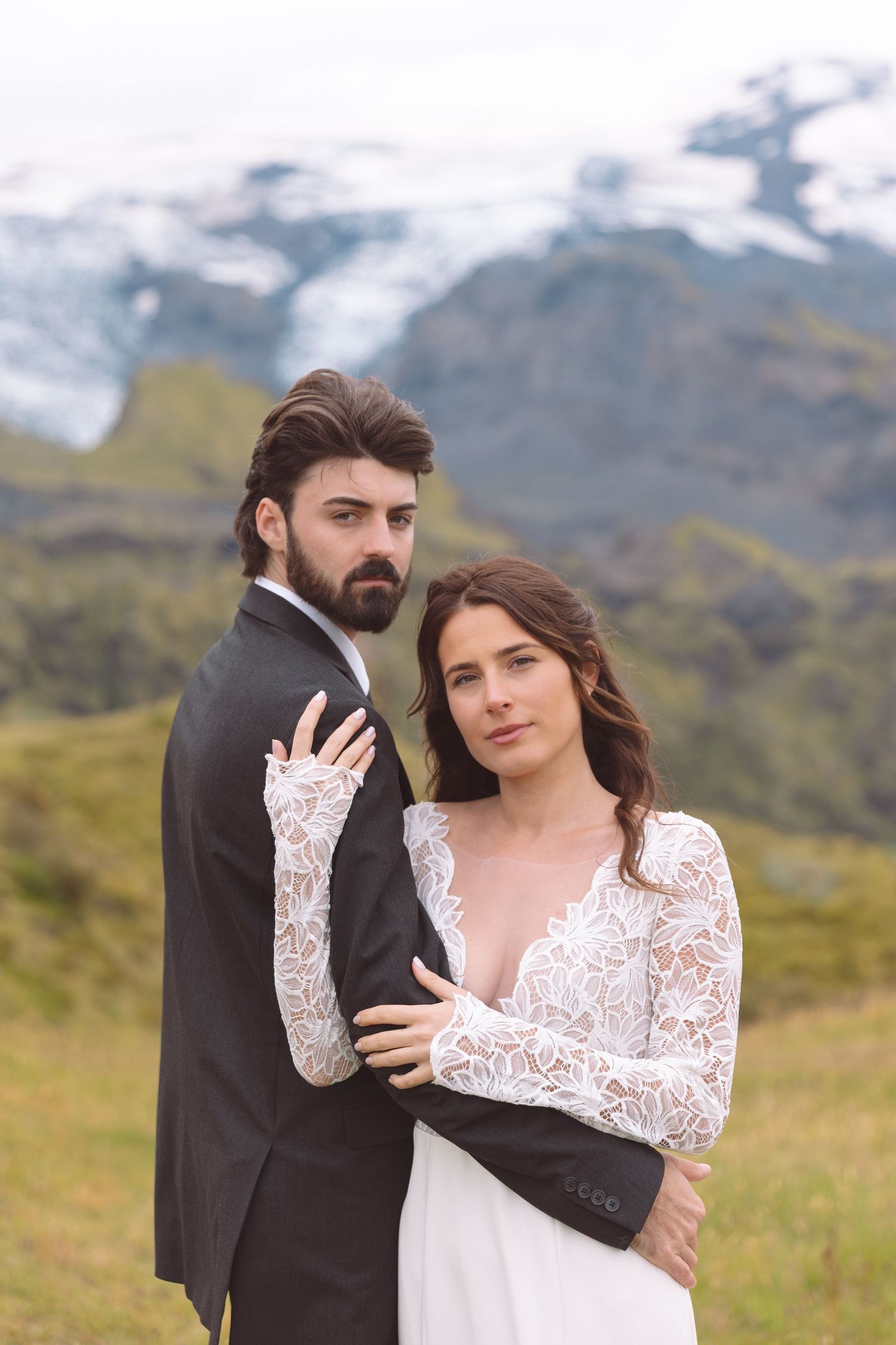 Bride in lace gown embraces bearded groom in dark suit with snowy mountains backdrop.