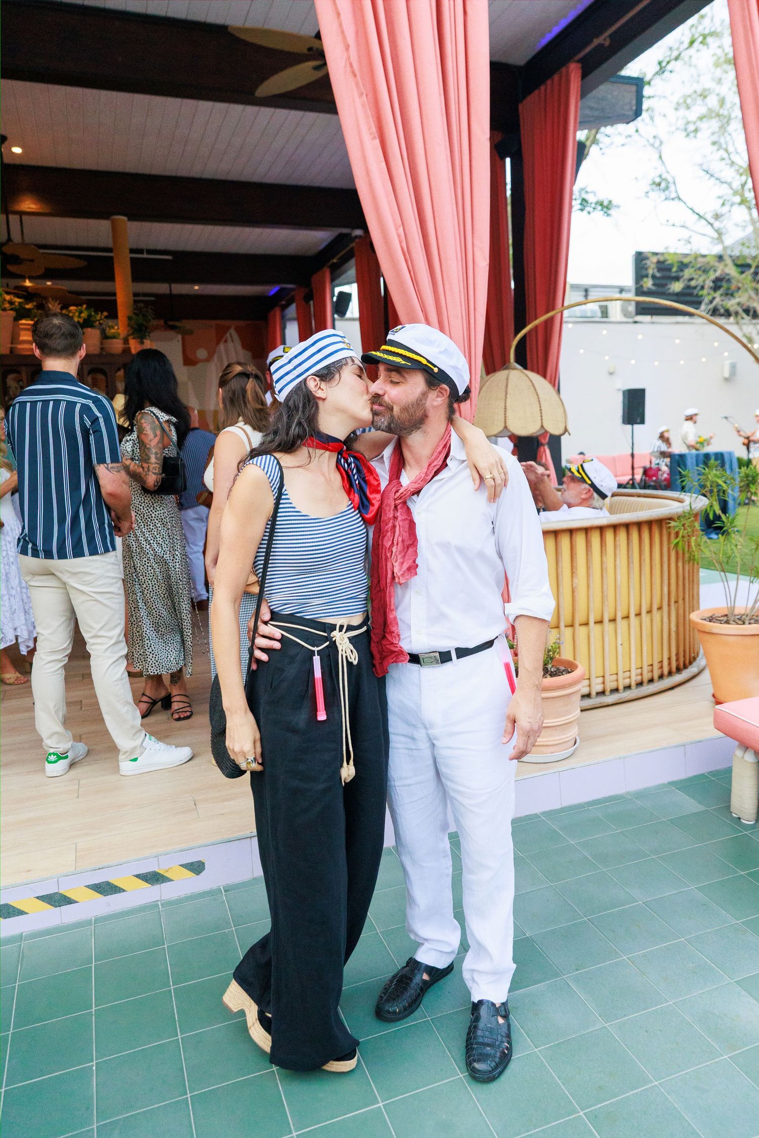 Couple in matching sailor outfits kissing at a festive outdoor party venue with pink curtains.