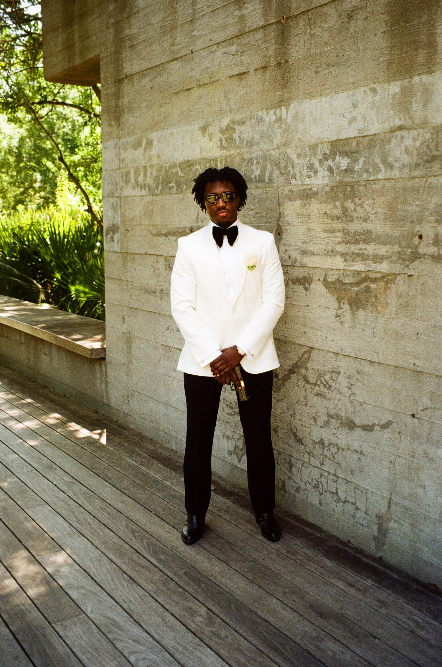 A young man in a white tuxedo jacket and black pants stands on a wooden deck beside a concrete wall.
