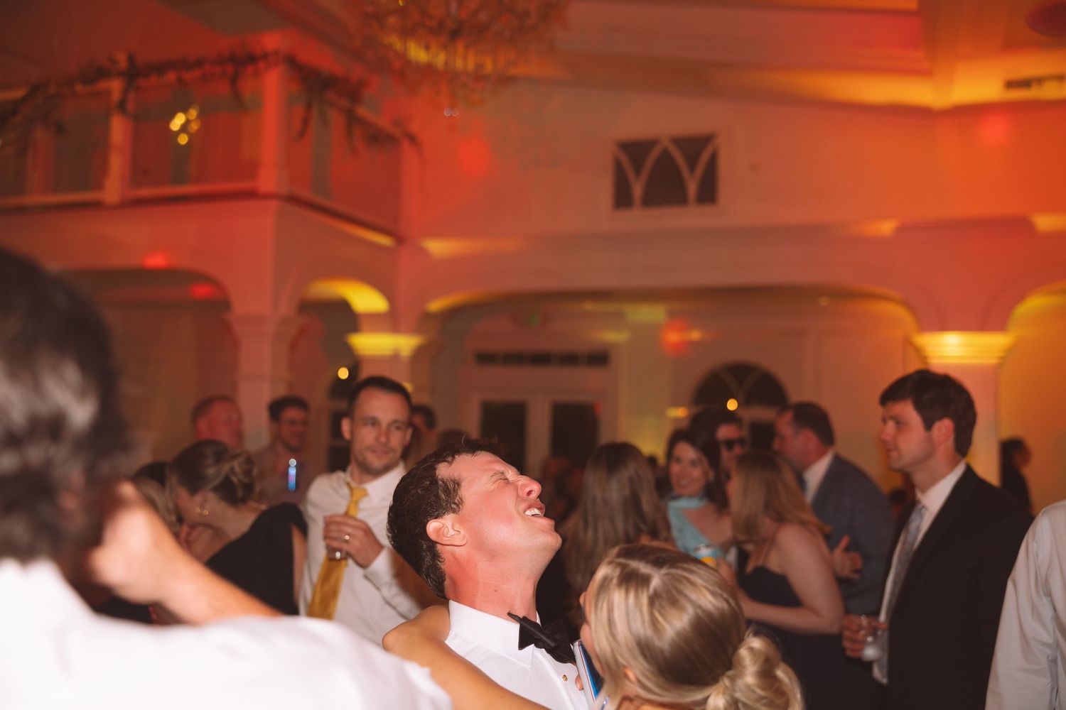Groom laughing joyfully surrounded by guests at a warmly lit wedding reception venue.