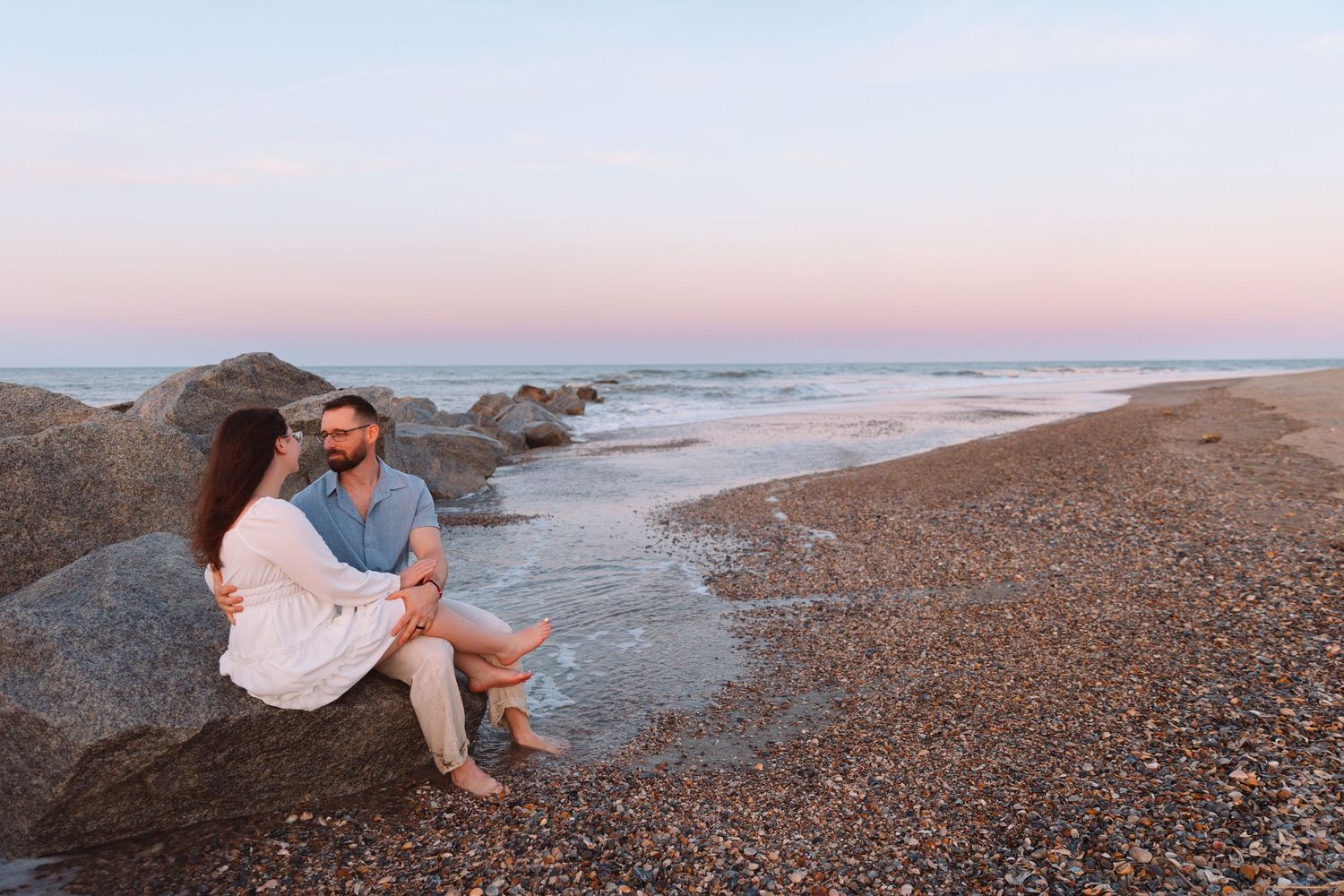 Couple sitting on rocks at a sandy beach during sunset, embracing with calm ocean waves in the background.
