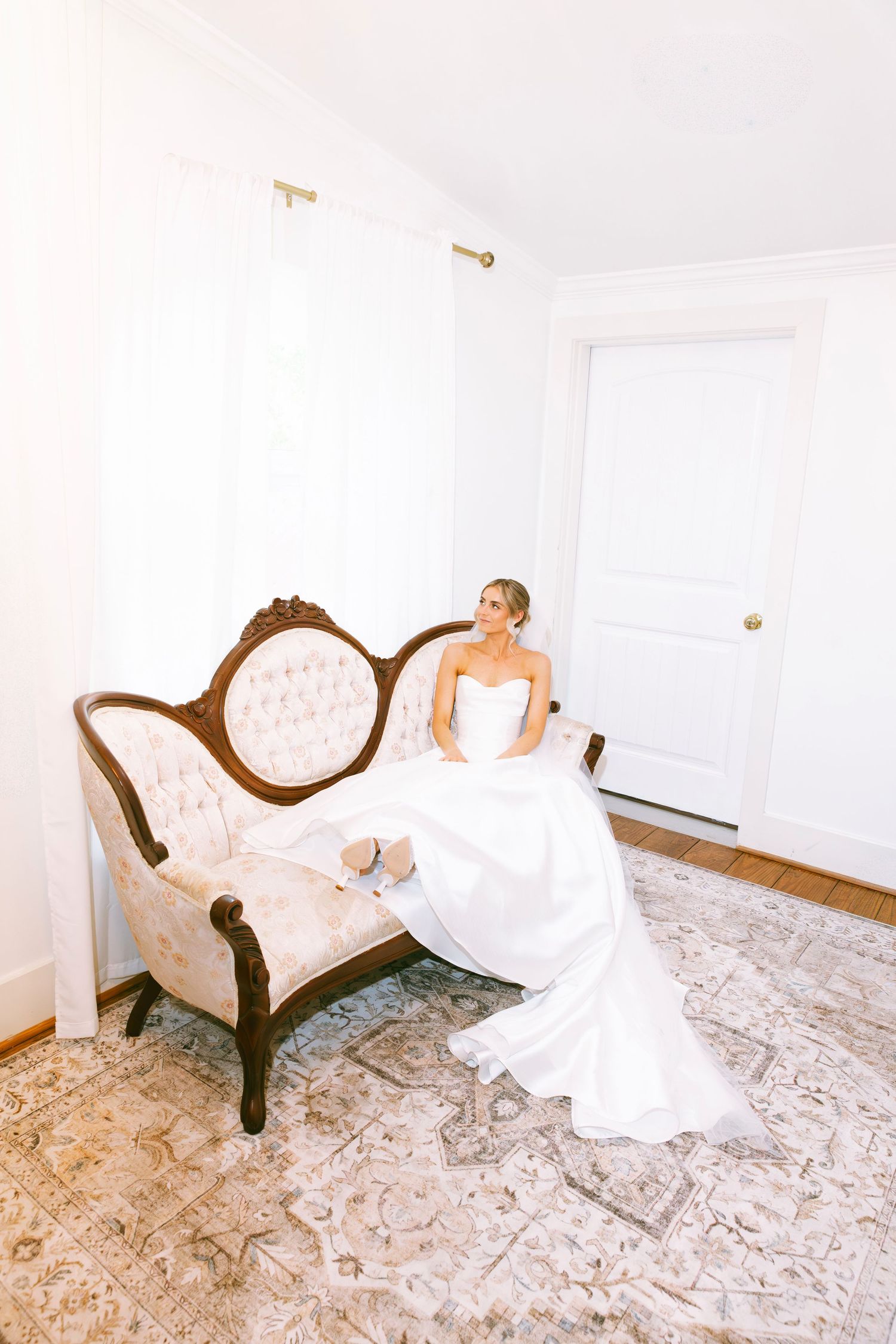 Bride in strapless white gown sits gracefully on vintage carved sofa in bright airy bridal suite.