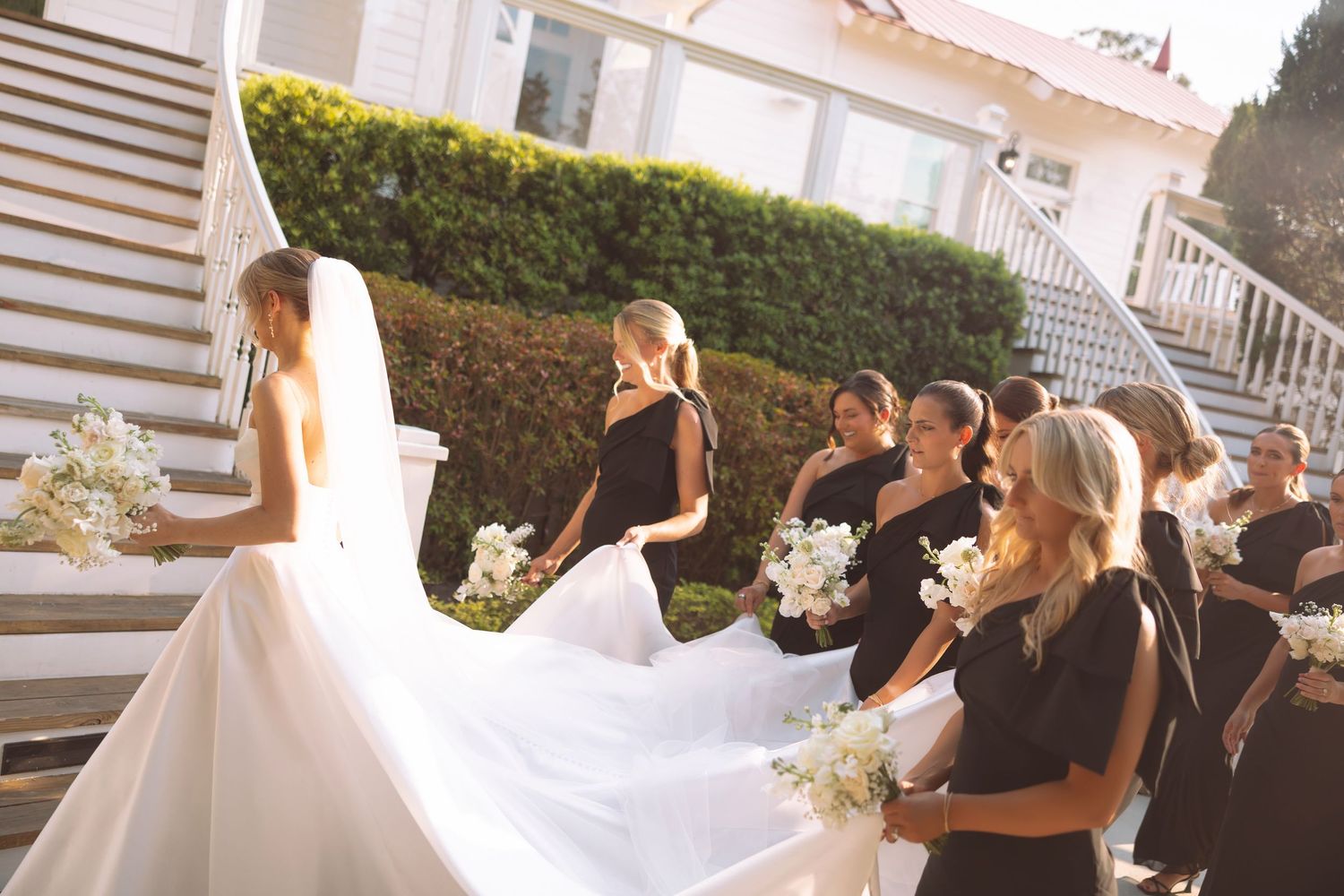 Bride in white gown with veil and bridesmaids in black dresses holding white bouquets on outdoor staircase.