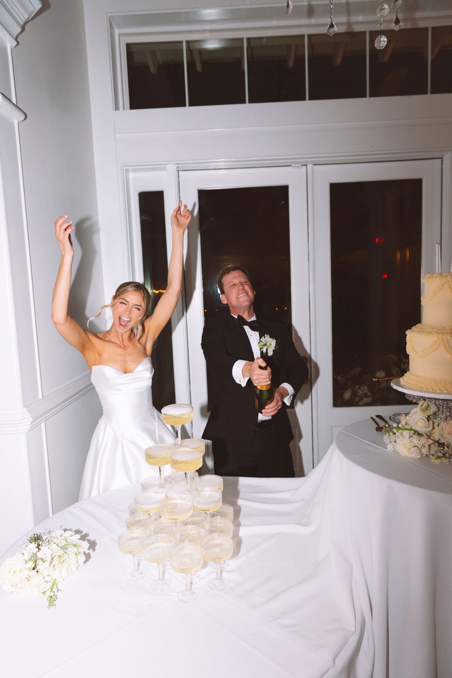 Bride and groom celebrating at wedding reception, popping champagne over a tower of glasses beside a tiered cake.