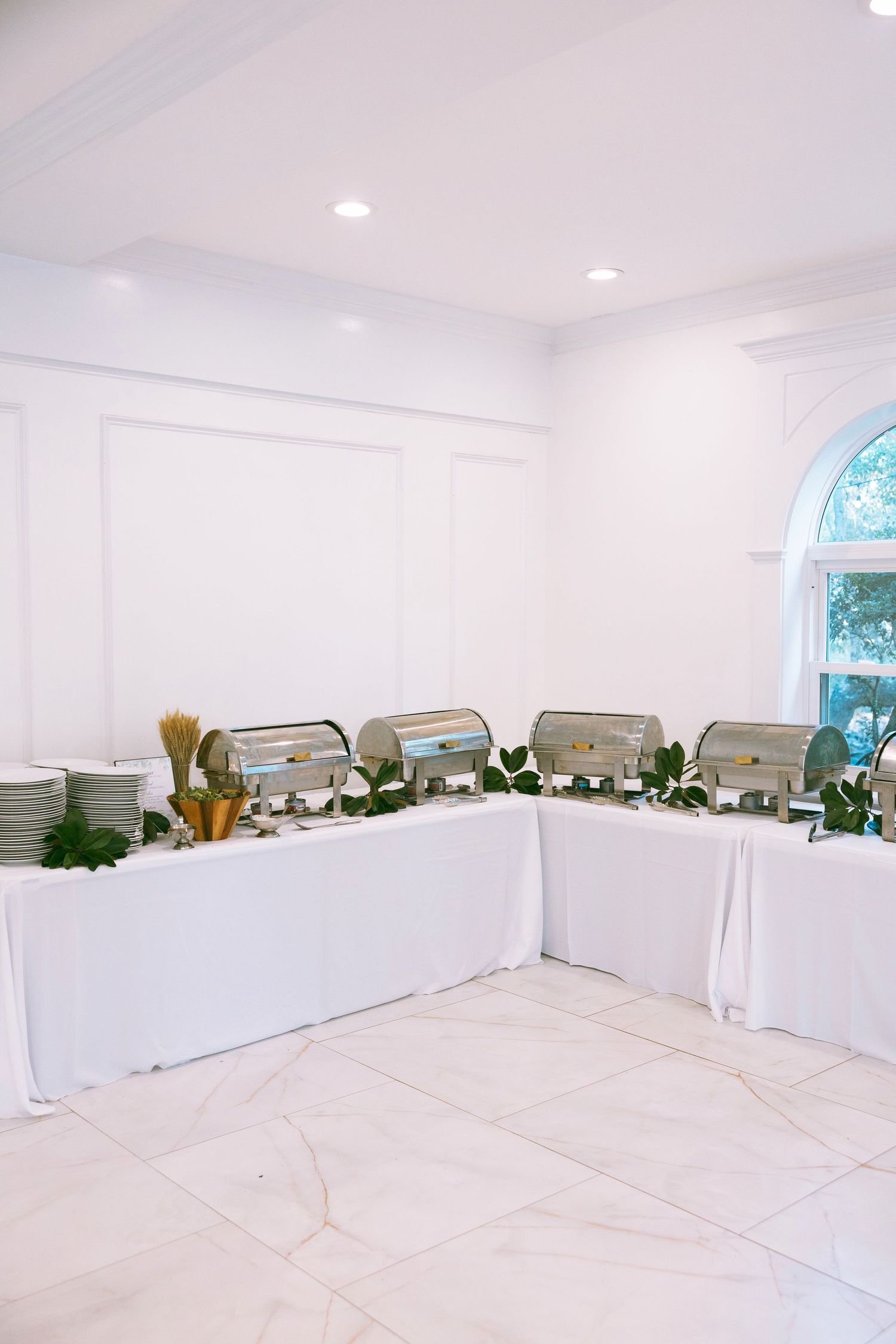 Elegant buffet setup with chafing dishes and greenery on white-draped tables in a bright, white-walled event hall.