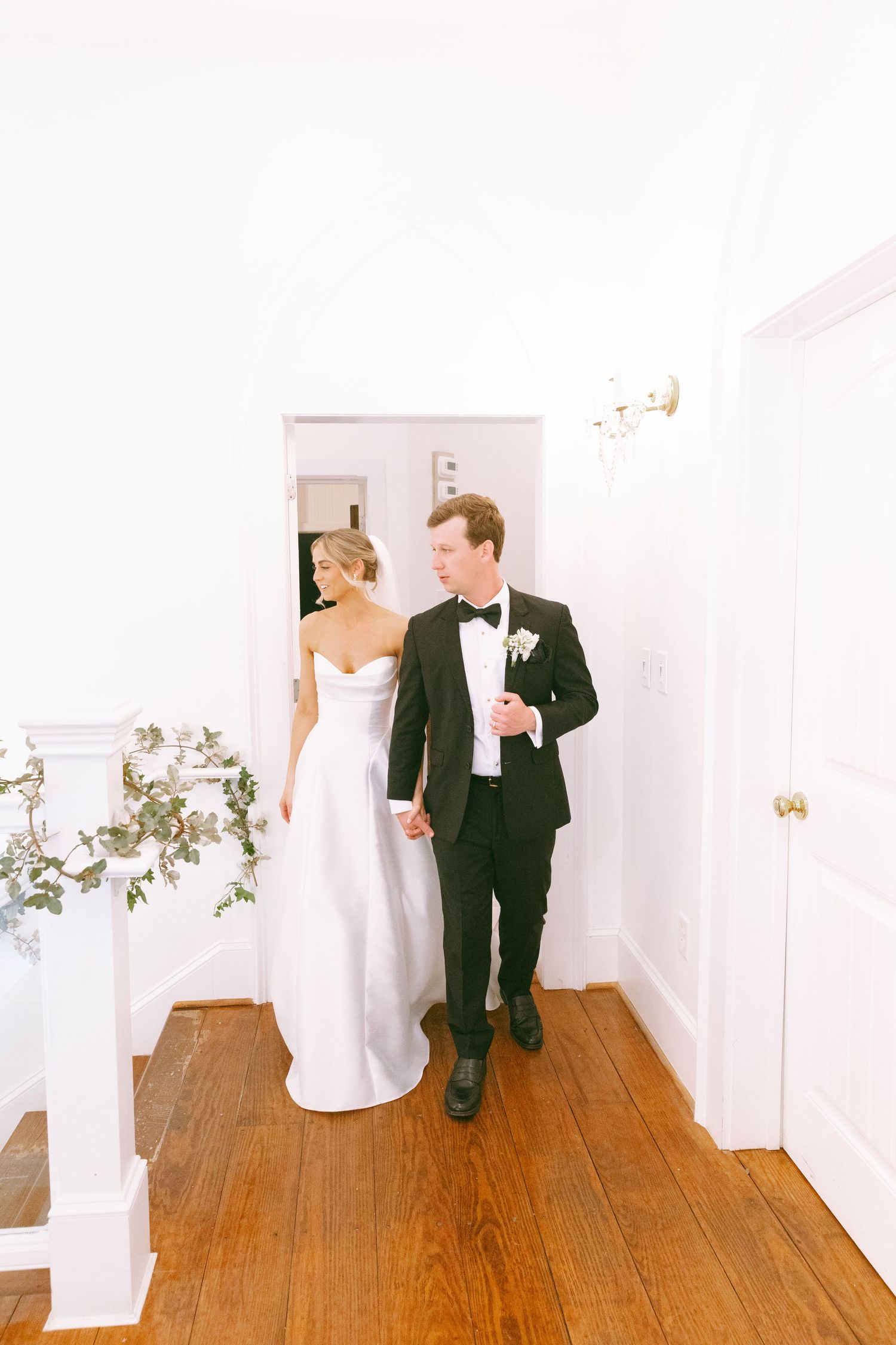 Bride in white gown and groom in black tuxedo walking hand in hand through a bright white hallway with hardwood floors.