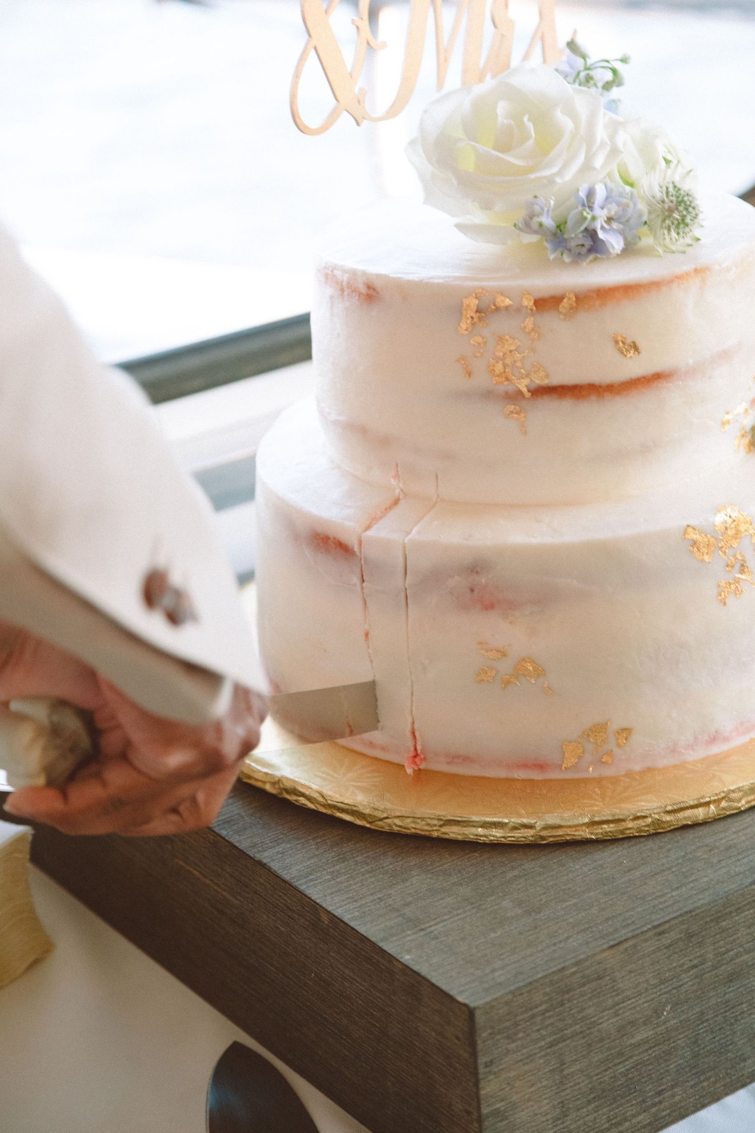 Close-up of two-tier naked wedding cake with gold leaf accents being sliced by bride and groom.