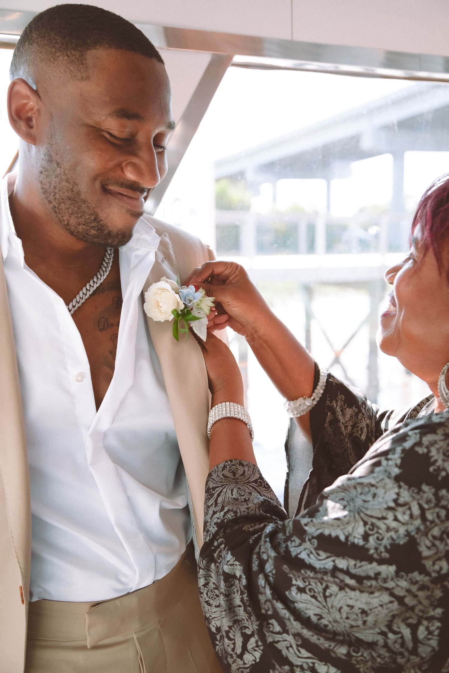 Woman pinning white floral boutonniere on smiling groom in white suit jacket before wedding.