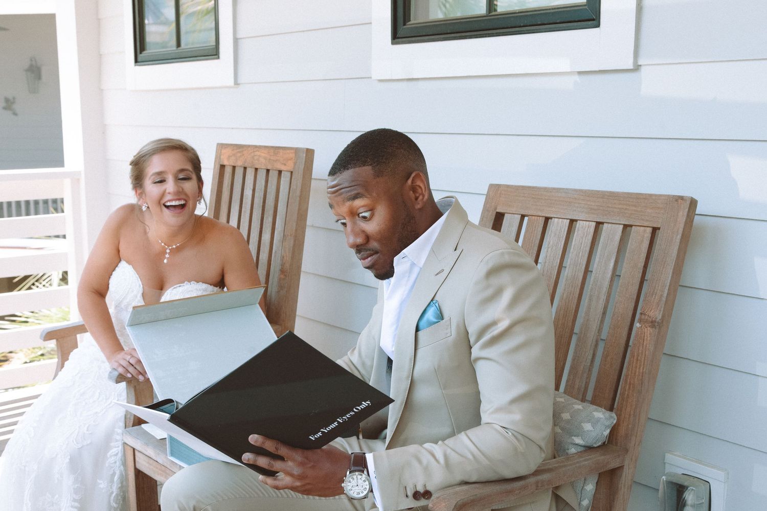 Bride laughing joyfully as groom opens wedding photo album while seated together on outdoor porch bench.