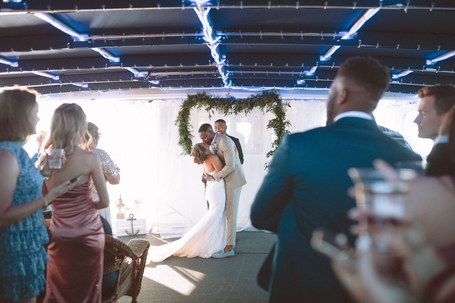 Bride and groom share first kiss under floral arch at intimate wedding ceremony with guests watching.