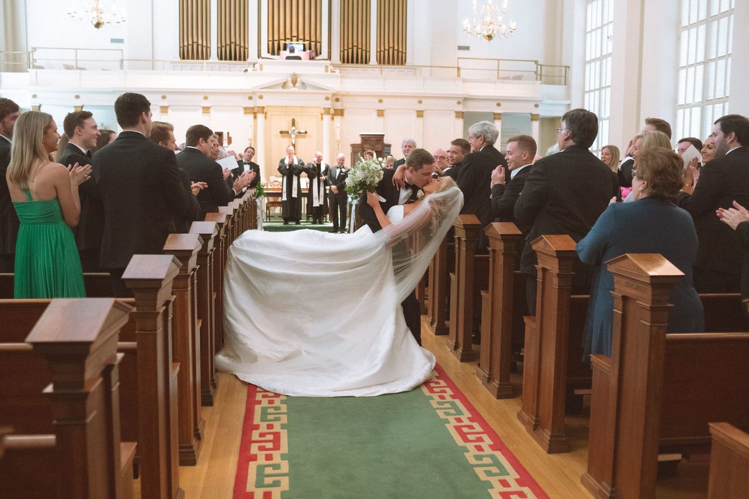 Bride and groom share their first kiss at the altar inside a classic white church with wooden pews.