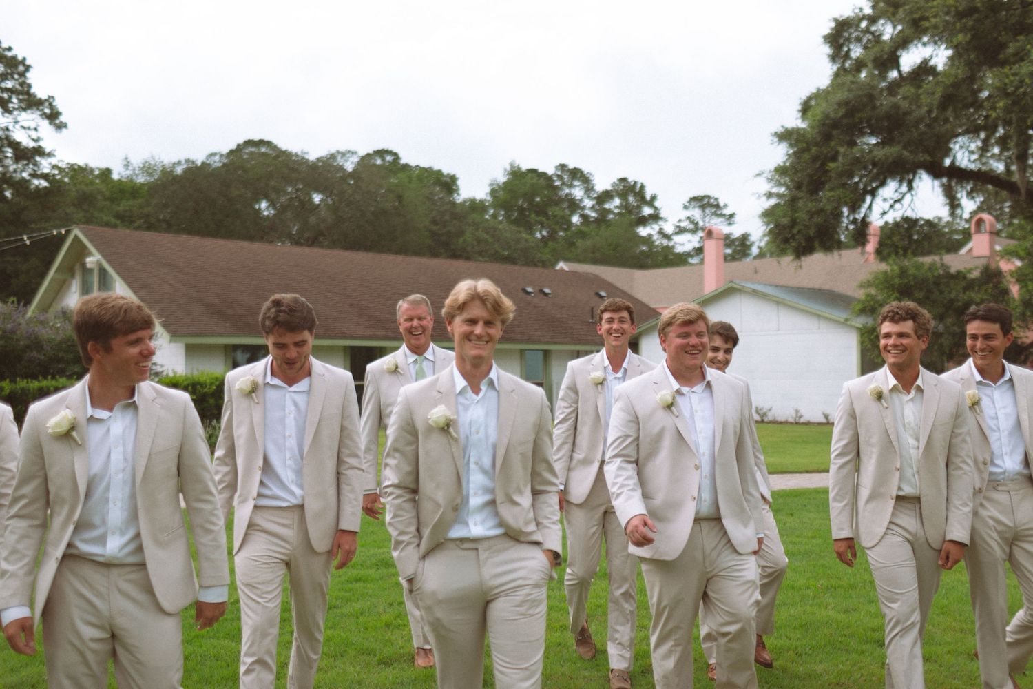 Groom and groomsmen walk confidently in matching cream suits across a green lawn.